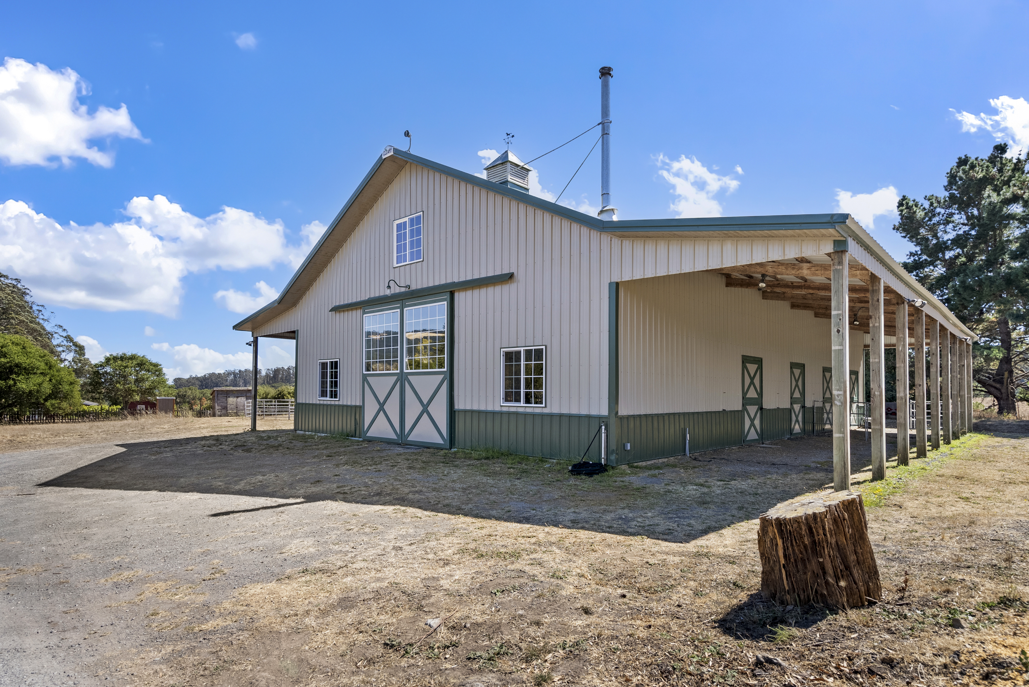 This image showcases a well-maintained, large metal barn building featuring a classic utilitarian design with green and beige siding. It includes a covered walkway or shed extension along the side, highlighting multiple stall-style doors, and displays traditional architectural elements like a cupola and a weather vane. The setting is a spacious, rural property grounds, emphasizing a functional space ideal for equestrian use or agricultural storage.