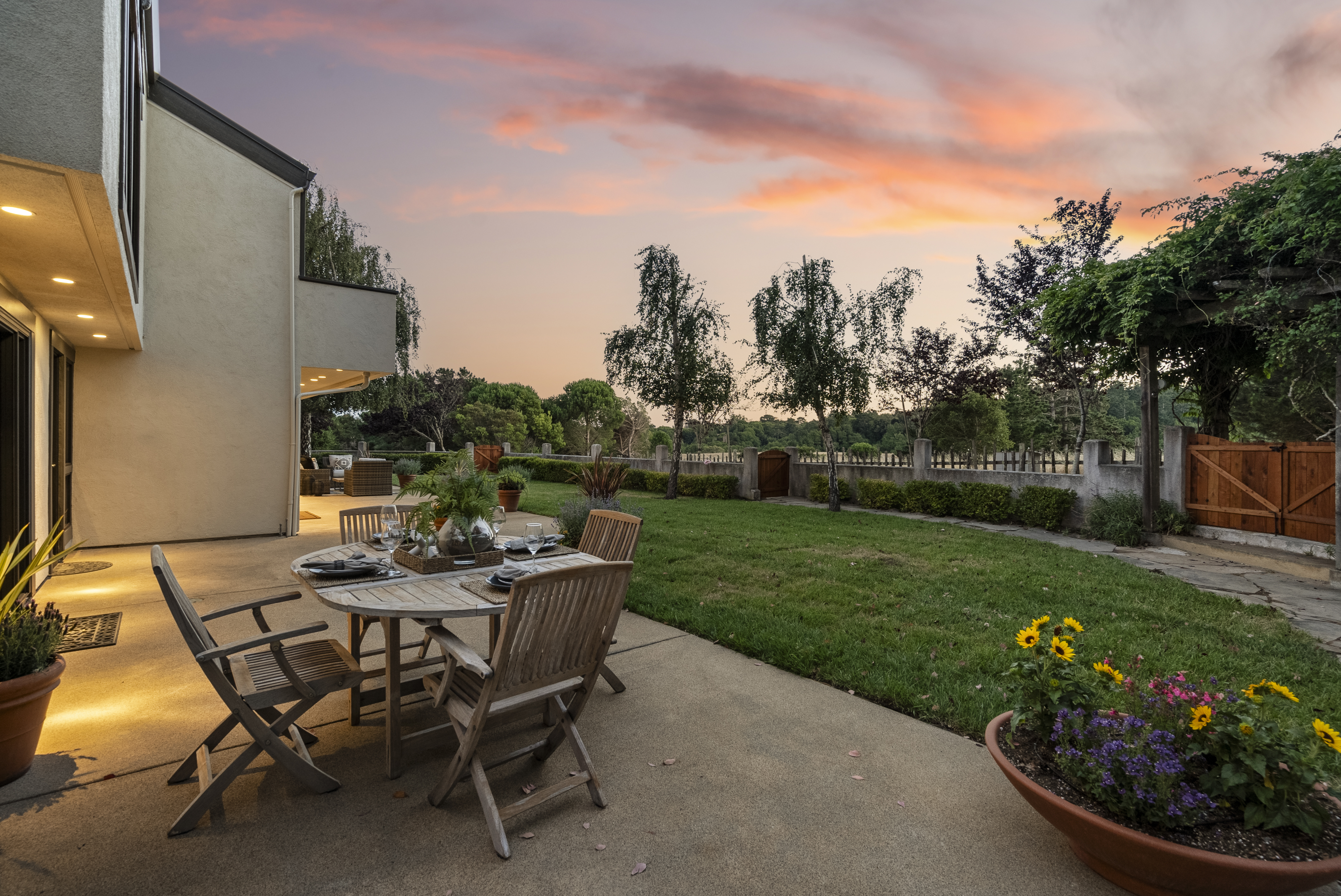 This beautiful outdoor living space features a spacious concrete patio set for dining at sunset with expansive views of the manicured lawn and surrounding greenery. The area is highlighted by a tasteful wooden table and chair set, potted plants, and warm ambient lighting from the home's exterior. It is an ideal setting for relaxed alfresco dining and garden entertaining.