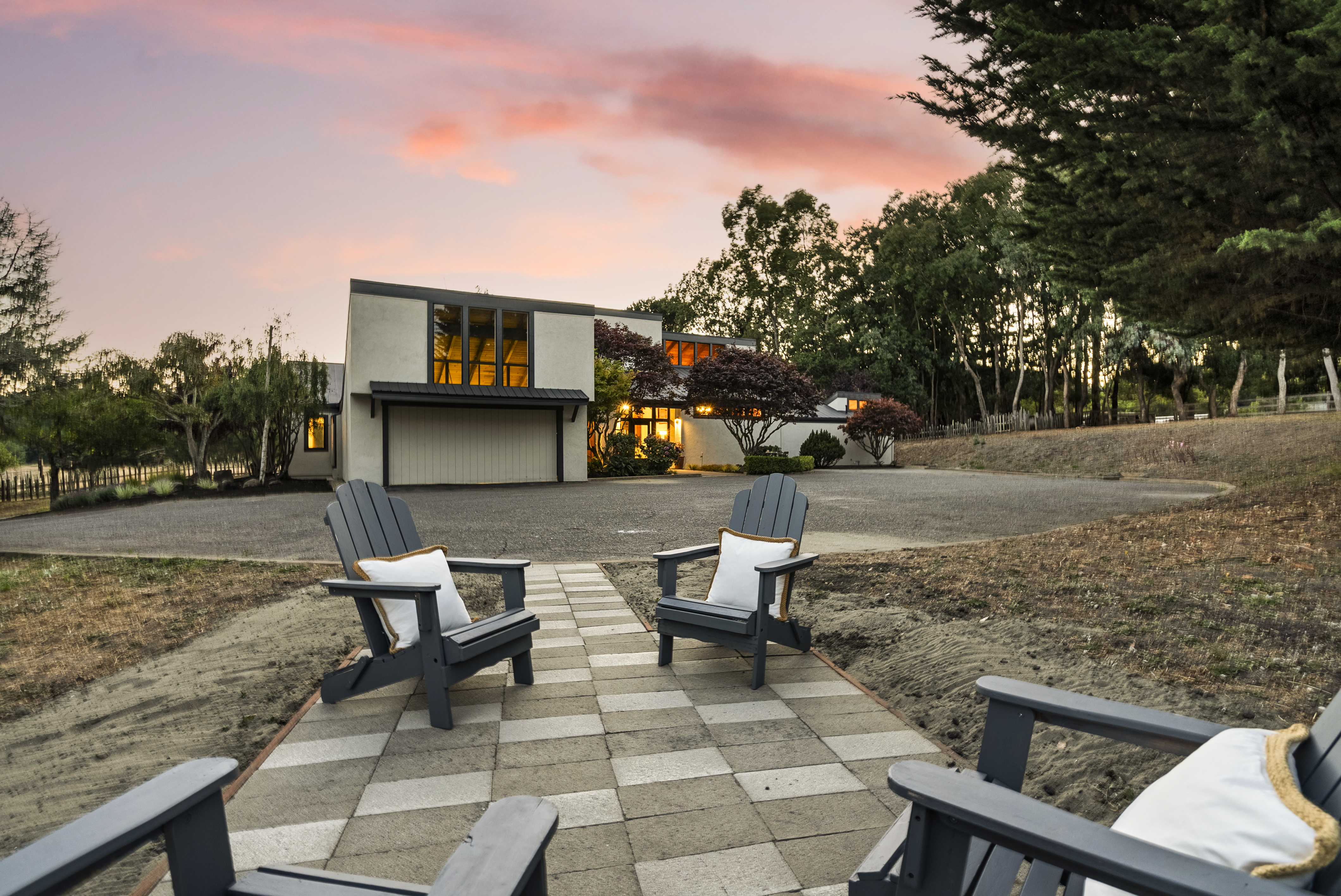This stunning contemporary home features a unique architectural design with a prominent front-facing garage and large, warm-lit windows. The inviting outdoor space includes a paved stone pathway lined with comfortable Adirondack chairs, perfect for enjoying the picturesque sunset over the landscape. Surrounded by mature trees and a natural setting, the house offers a sophisticated yet serene curb appeal.