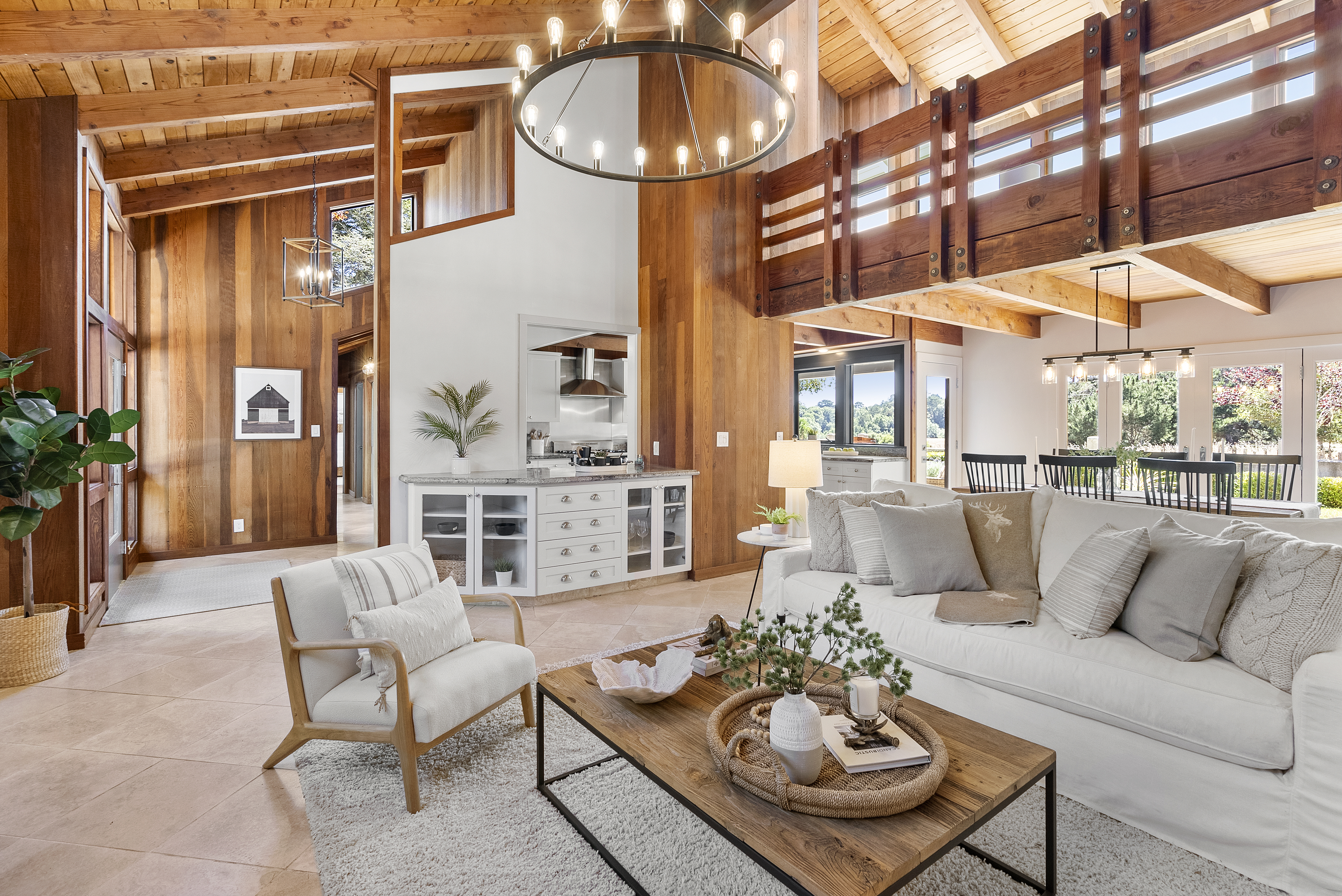 This spacious, open-concept living room features soaring wood-beamed ceilings and warm wood-paneled walls, creating a cozy yet airy mountain-lodge aesthetic. The design includes a contemporary circular chandelier, a comfortable white sofa with throw pillows, and a rustic wood coffee table, all centered around a well-lit living space. Neutral-toned tile floors flow seamlessly into the adjacent kitchenette, emphasizing a bright and inviting atmosphere.