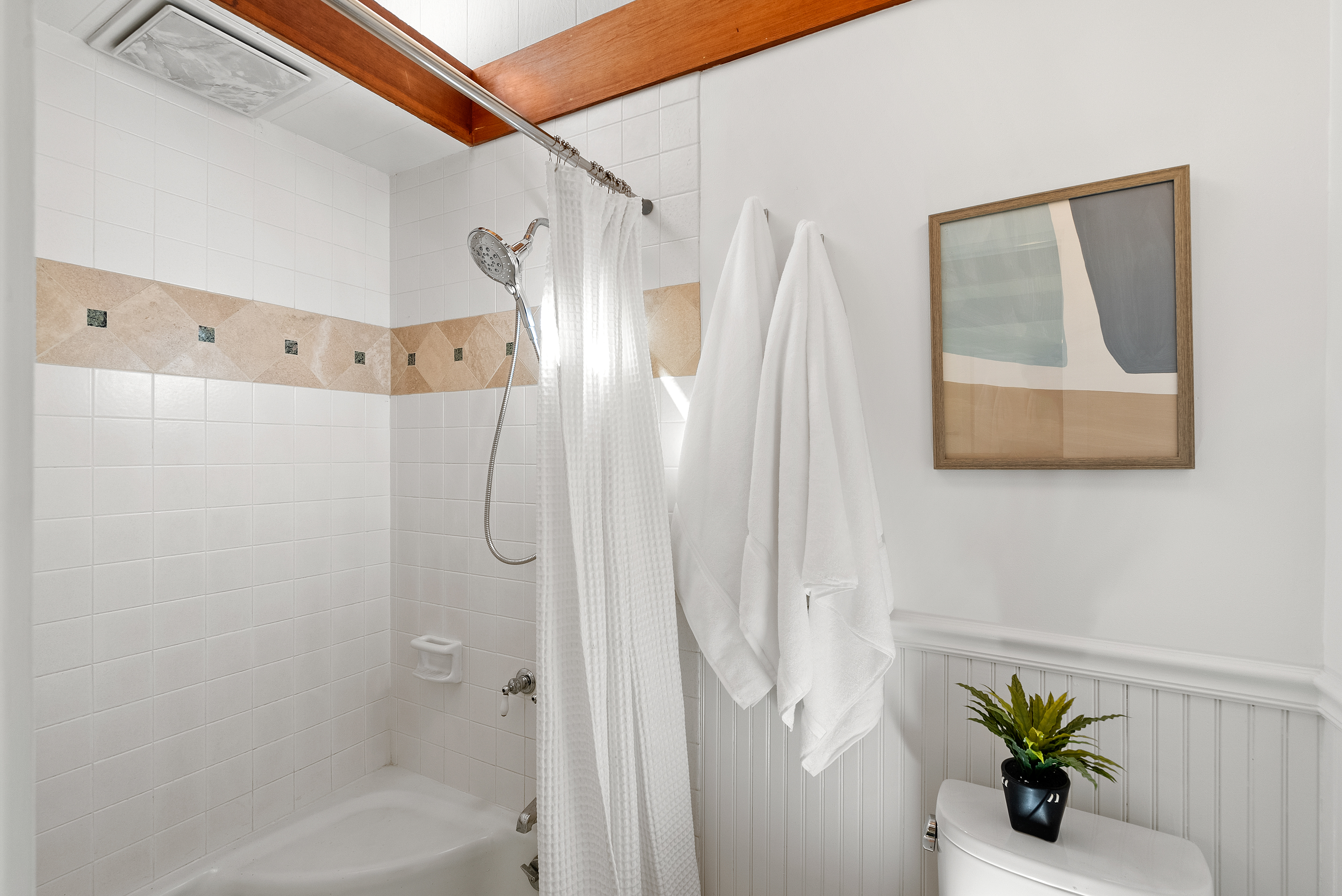 This clean and bright bathroom features a combined shower and tub with simple white subway tiling and a beige decorative accent band. A white shower curtain, dual white hanging towels, and a small potted plant on the toilet tank add a fresh, inviting touch to the space. The room is finished with light-colored wainscoting and a wooden framed abstract art piece, creating a minimalist yet homey atmosphere.