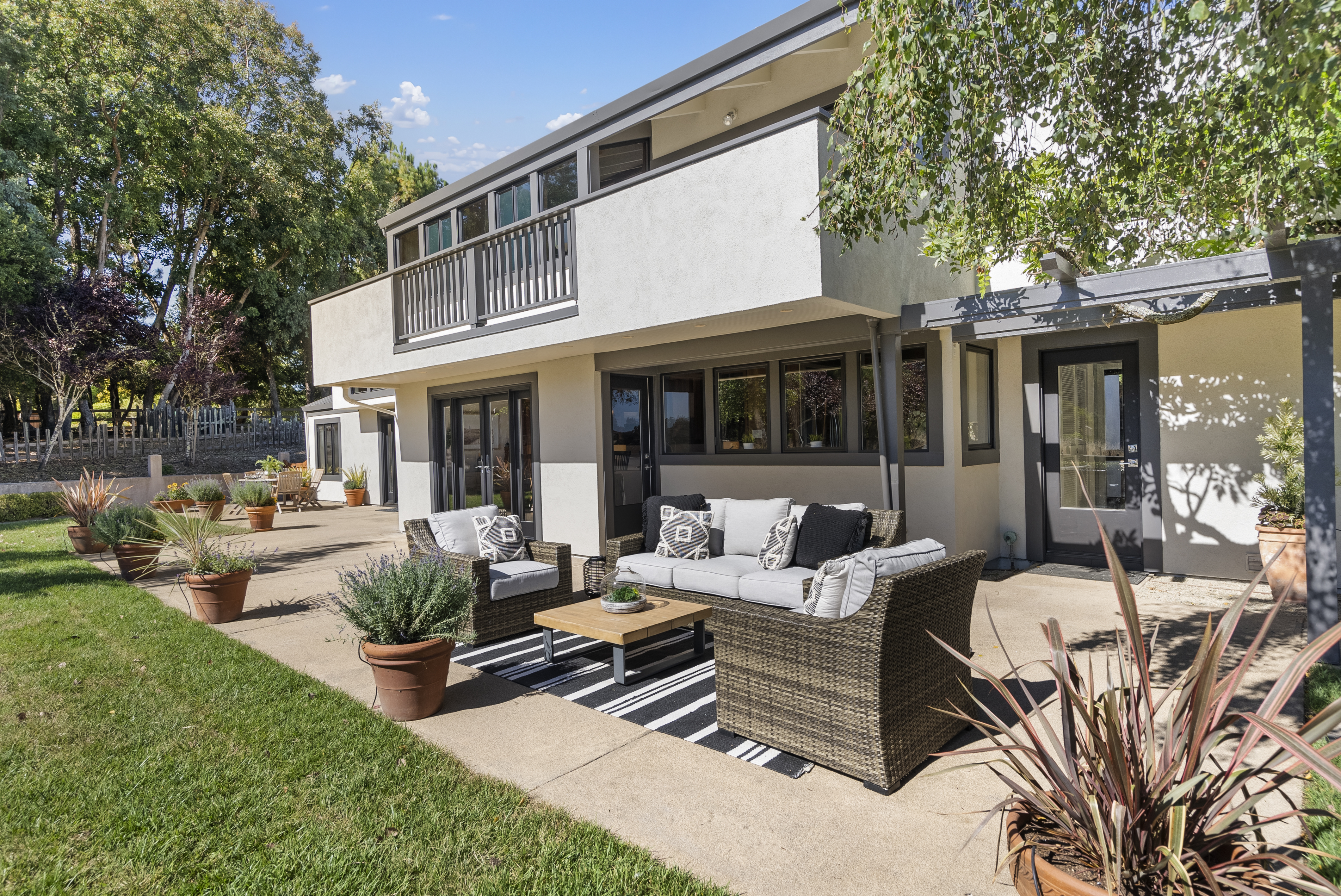 This modern multi-level residence features a spacious rear patio equipped with comfortable outdoor wicker furniture and a striped area rug. The exterior showcases clean lines, neutral stucco walls, and a recessed balcony, seamlessly blending indoor-outdoor living. Lush landscaping in terracotta pots and a well-manicured lawn create an inviting and relaxing environment for entertaining.