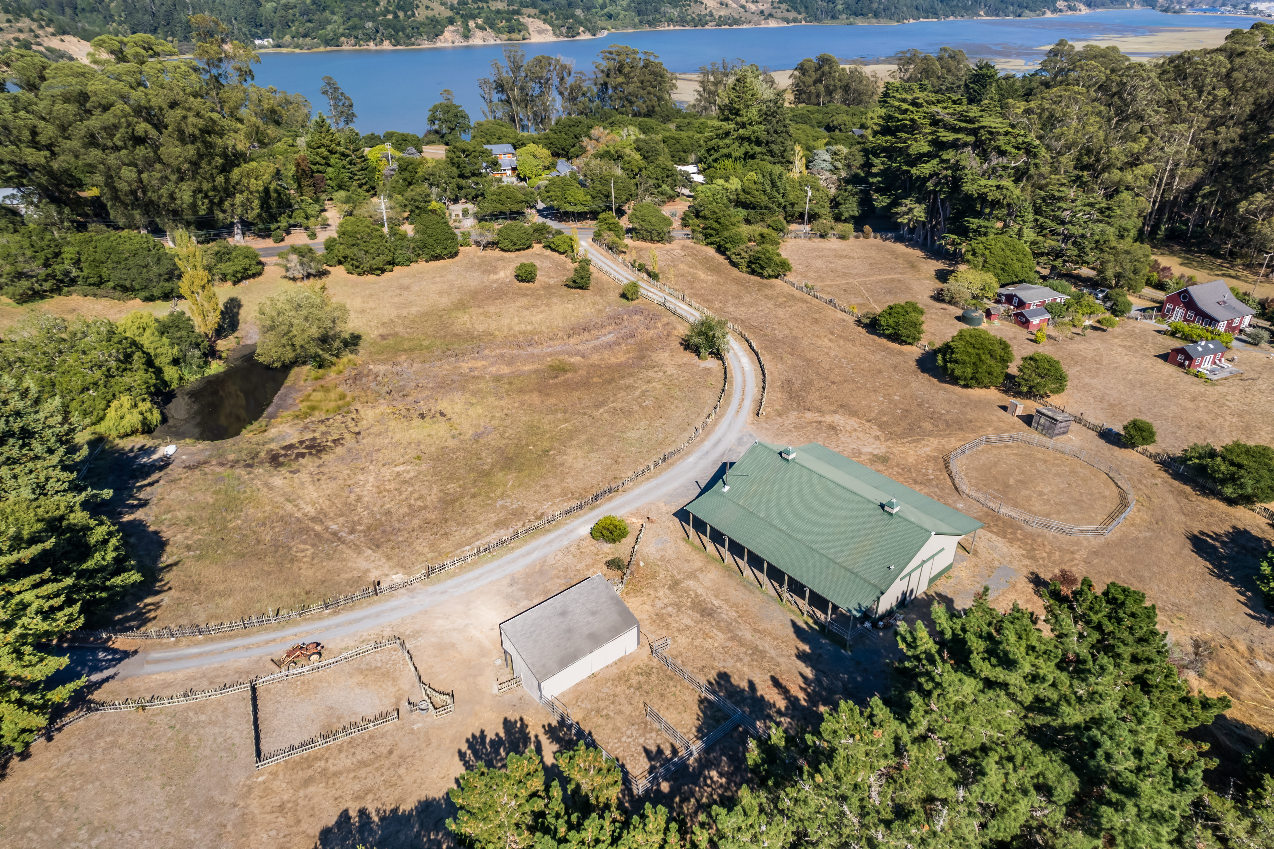 This stunning high-angle aerial view showcases a sprawling rural property set against a scenic backdrop of a body of water and rolling hills. The land features a large green-roofed barn, a secondary outbuilding, fenced equestrian rings, and a winding driveway that leads through the dry, open fields. The generous acreage, rustic charm, and peaceful surroundings define this property as a fantastic retreat with significant potential for livestock or hobby farming.