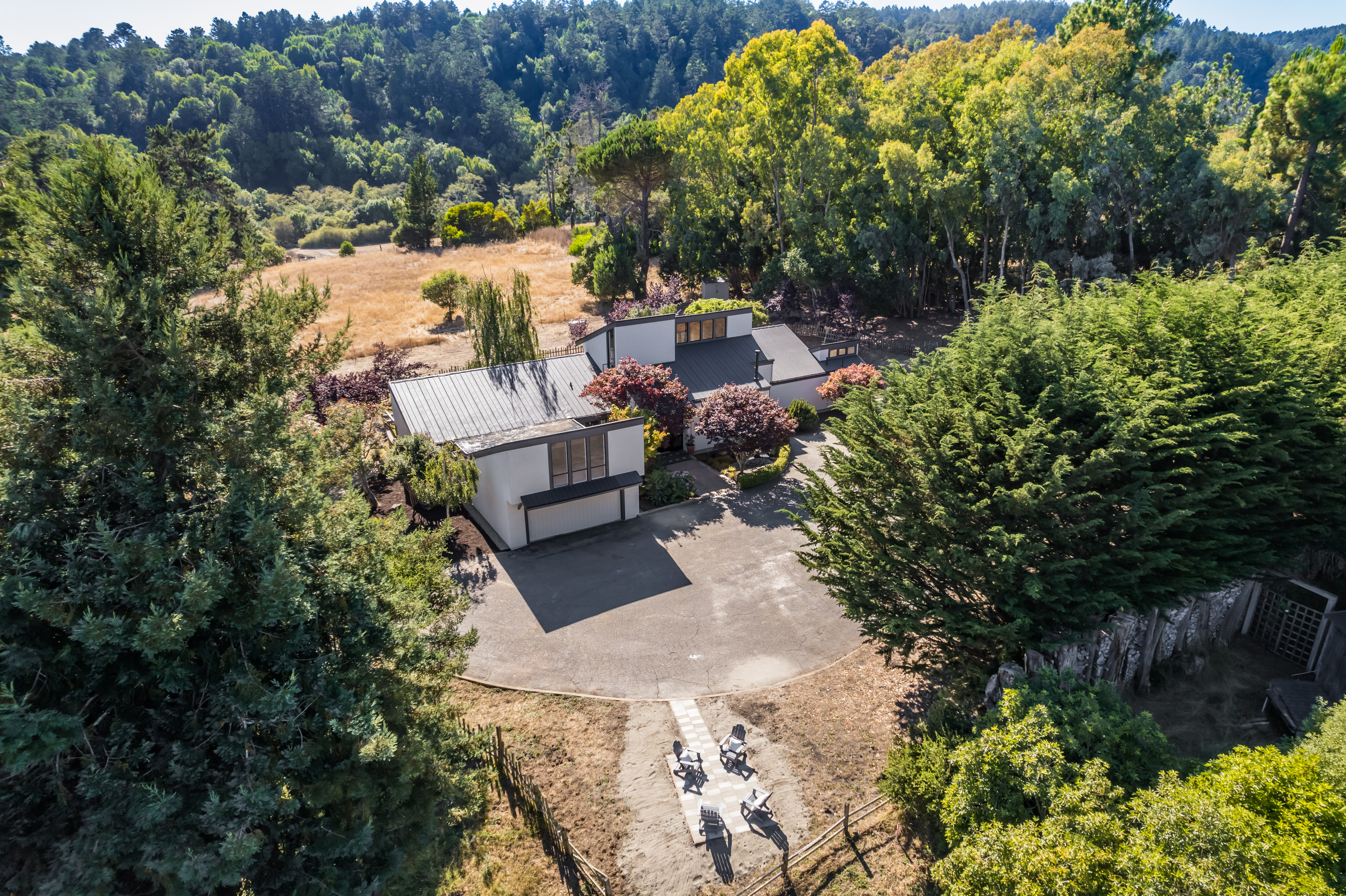 This stunning aerial view captures a contemporary, multi-level home nestled within a tranquil wooded landscape. The structure features modern clean lines, a metal roof, and a welcoming circular driveway that leads to a paved patio area equipped with comfortable outdoor seating. The surrounding dense forest and gentle hillside setting offer significant privacy and a peaceful, nature-integrated lifestyle.
