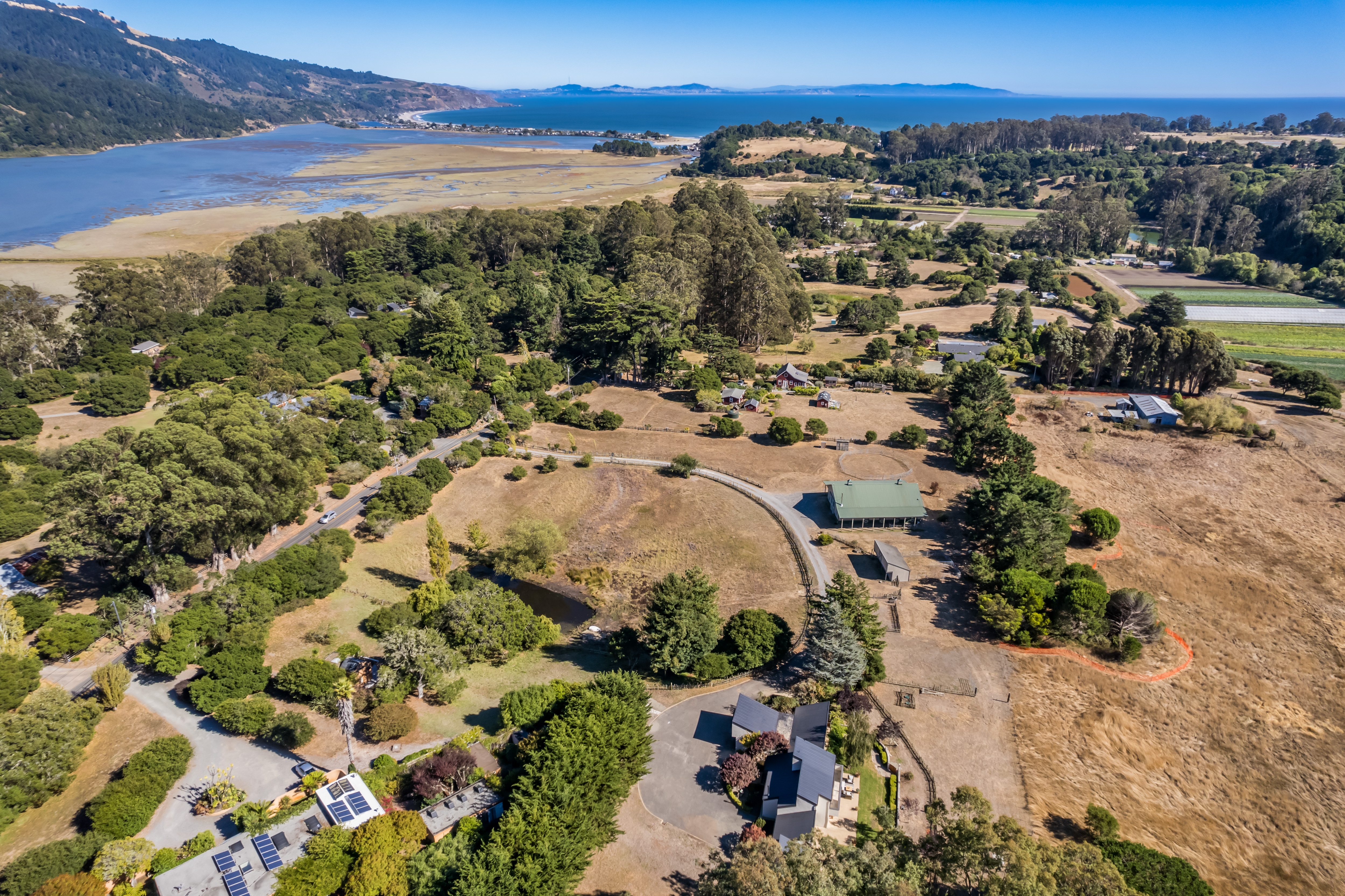 This panoramic aerial view captures a sprawling rural property featuring multiple residential structures surrounded by extensive natural landscapes. The scene highlights large wooded areas, open fields, and proximity to a coastal estuary and distant ocean views, emphasizing a serene, country lifestyle. A well-maintained driveway winds through the property, showcasing the significant acreage and peaceful, secluded setting ideal for a private estate.