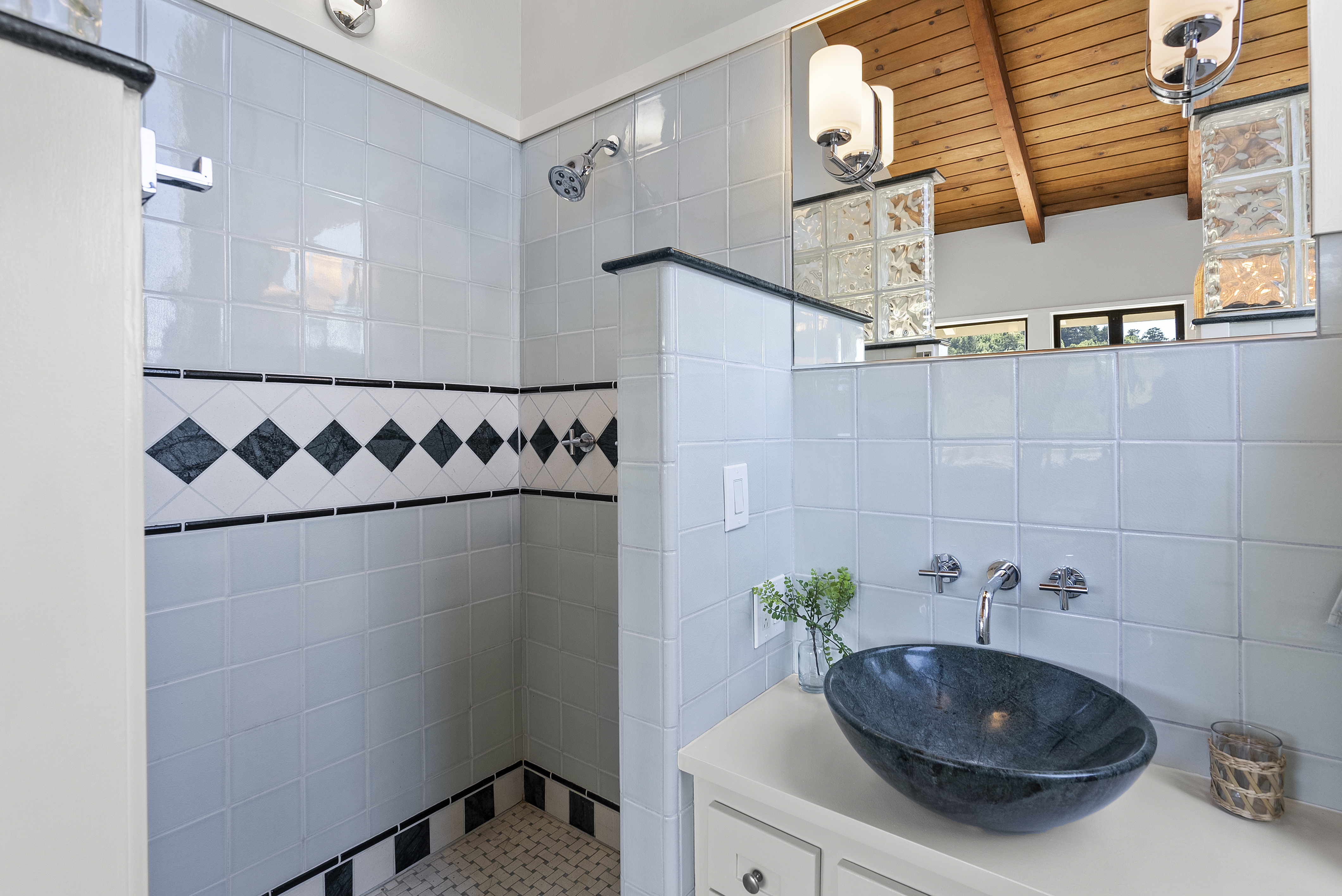 This bathroom features a contemporary design highlighted by a distinctive stone vessel sink and wall-mounted chrome fixtures. Pale blue square tiling covers the walls, accented by a decorative black-and-white diamond border, while a wood-beamed ceiling adds warmth. The layout includes a semi-enclosed shower area and a glass block element, creating a unique and stylish aesthetic.