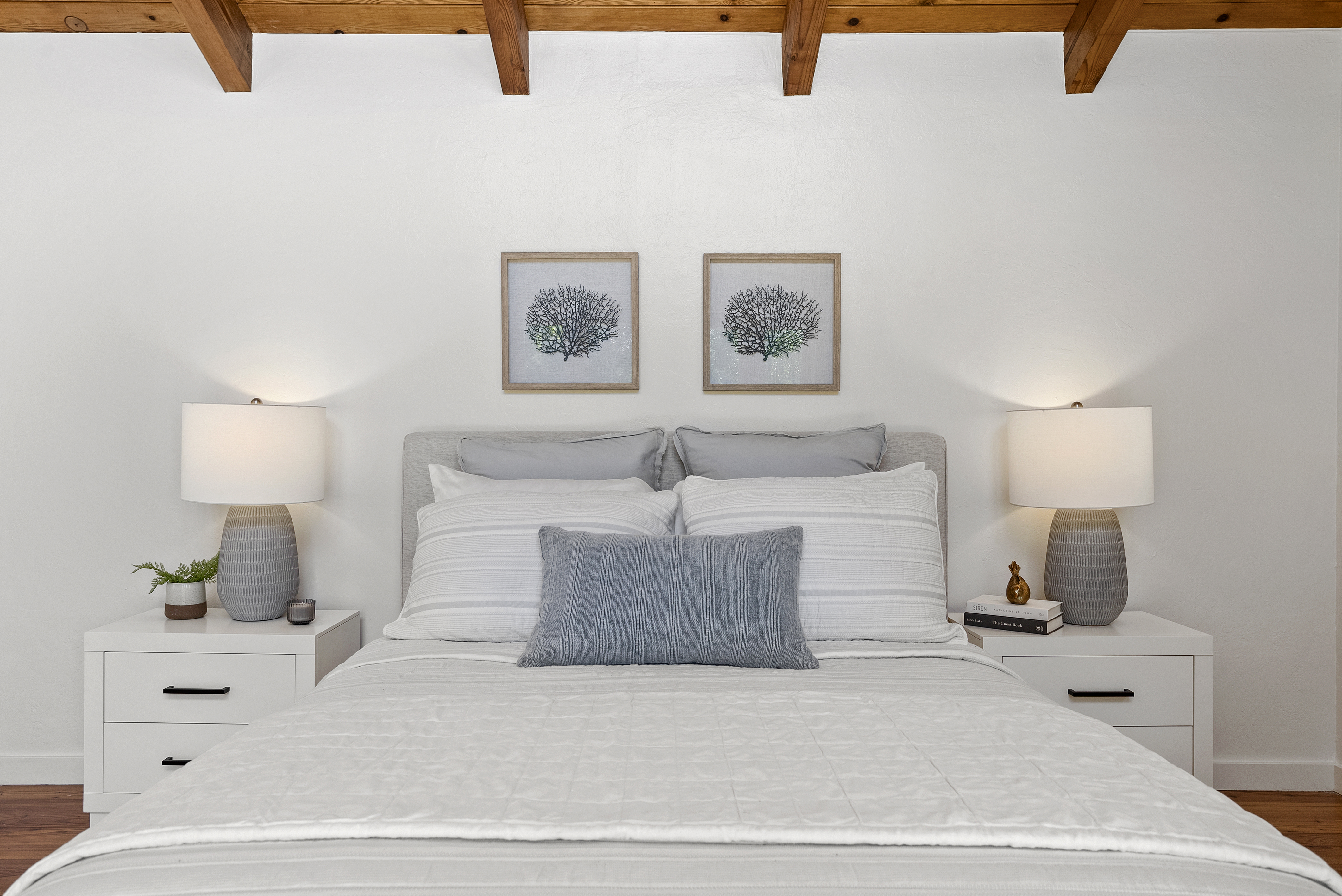 This serene primary bedroom features a clean, neutral color palette with sophisticated coastal-inspired accents. The space includes a pair of stylish gray textured lamps, matching bedside tables, and subtle wall art, all framed by elegant exposed wood ceiling beams. The composition is balanced and inviting, highlighting a comfortable, well-staged retreat.