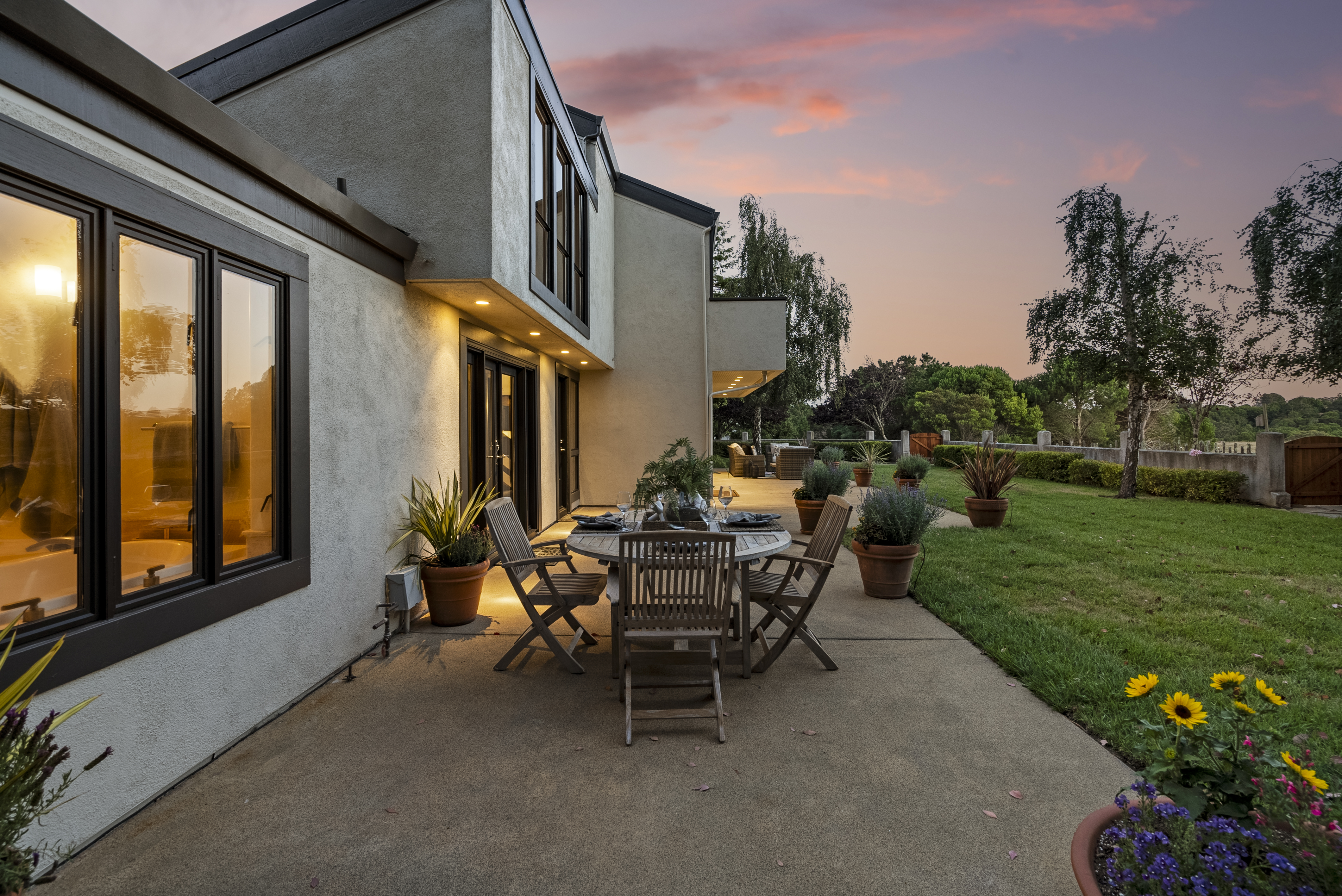 This image showcases an inviting rear patio area featuring a functional dining set, perfect for outdoor entertaining as the sun sets. The modern stucco home includes large windows providing a warm, interior glow, seamlessly connecting indoor and outdoor living spaces. Lush landscaping, potted plants, and a manicured lawn create a serene and well-maintained backyard environment.