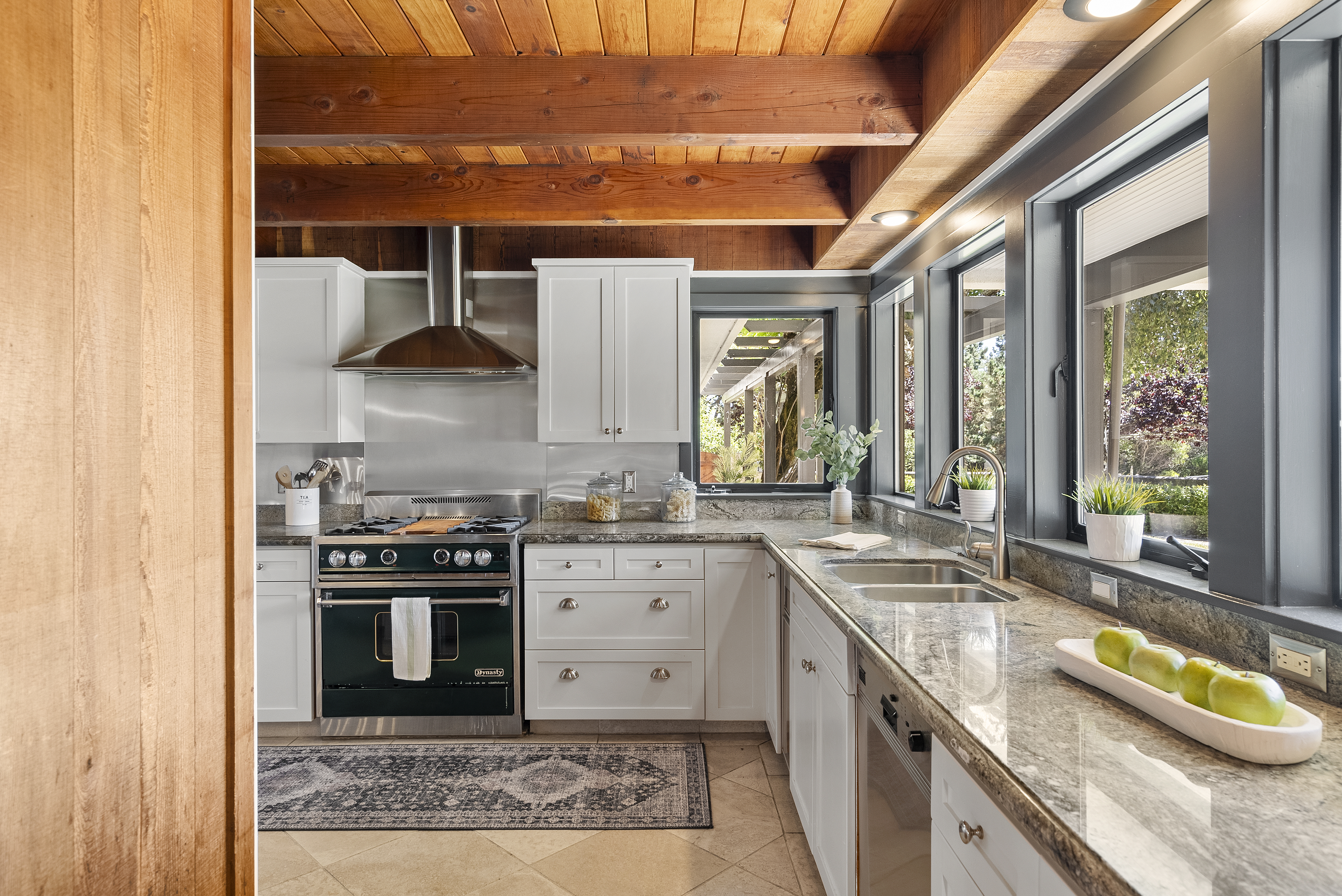 This kitchen features a warm, rustic aesthetic highlighted by exposed wooden beams on the vaulted ceiling and natural wood paneling. The space is well-equipped with modern white cabinetry, polished granite countertops, and a professional-grade dark green range with a stainless steel hood. Large windows fill the area with natural light, creating a bright and inviting environment perfect for cooking and entertaining.