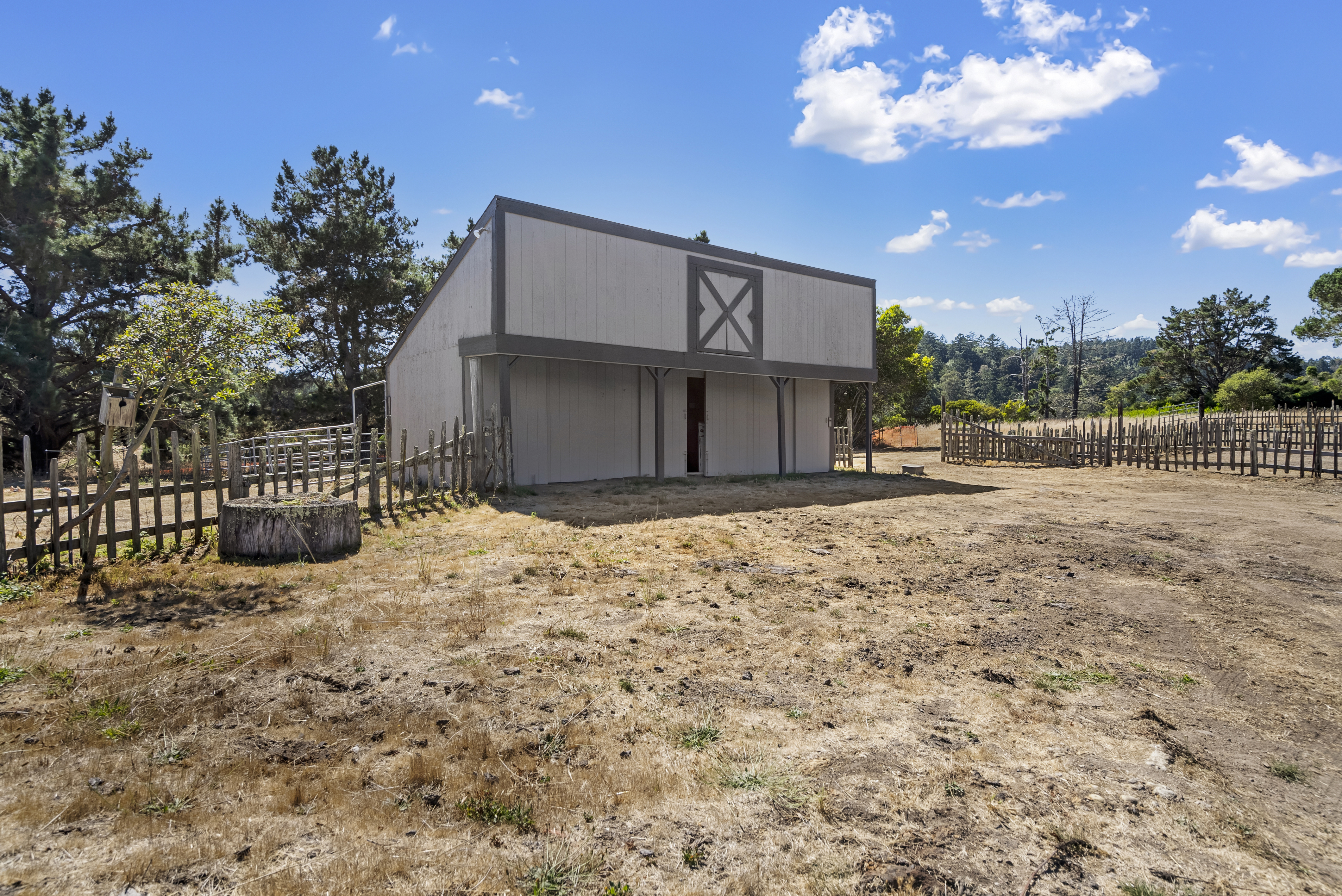 This image features a rustic, wood-sided barn or shed set in a dry, open field under a vibrant blue sky. The structure includes simple architectural detailing, such as a signature X-braced gable vent, and is partially enclosed by a weathered wooden picket fence. The setting suggests a rural property with ample space and natural charm, ideal for agricultural or hobby-farm use.