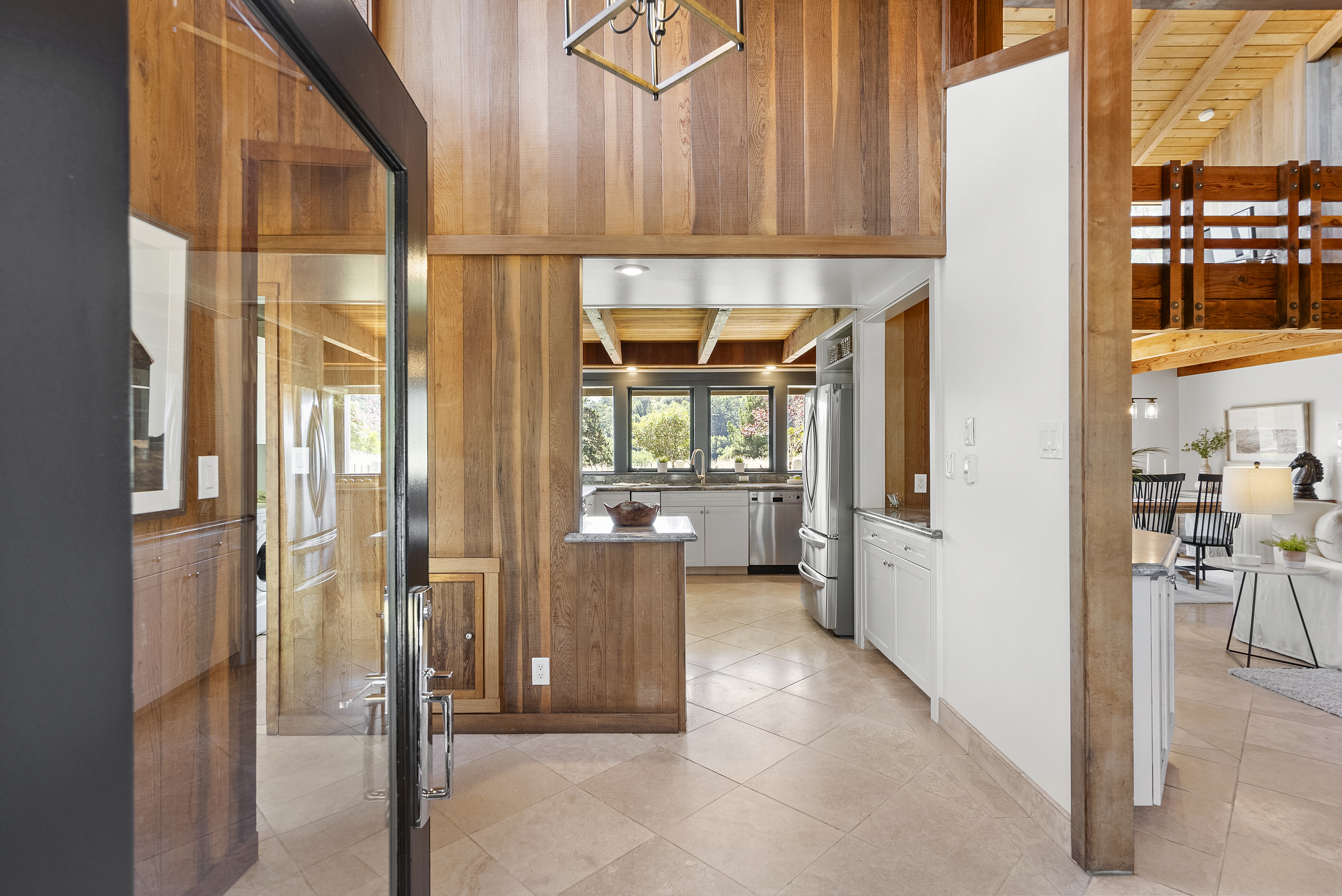 This interior view shows a kitchen transitioning from a wood-paneled entryway, highlighting a blend of rustic charm and modern functionality. The kitchen features stainless steel appliances, white cabinetry, and a clean tiled floor, while exposed wooden ceiling beams and paneling add warmth throughout the space. The open-concept layout creates an inviting flow, connecting the kitchen to a bright, comfortable living area.