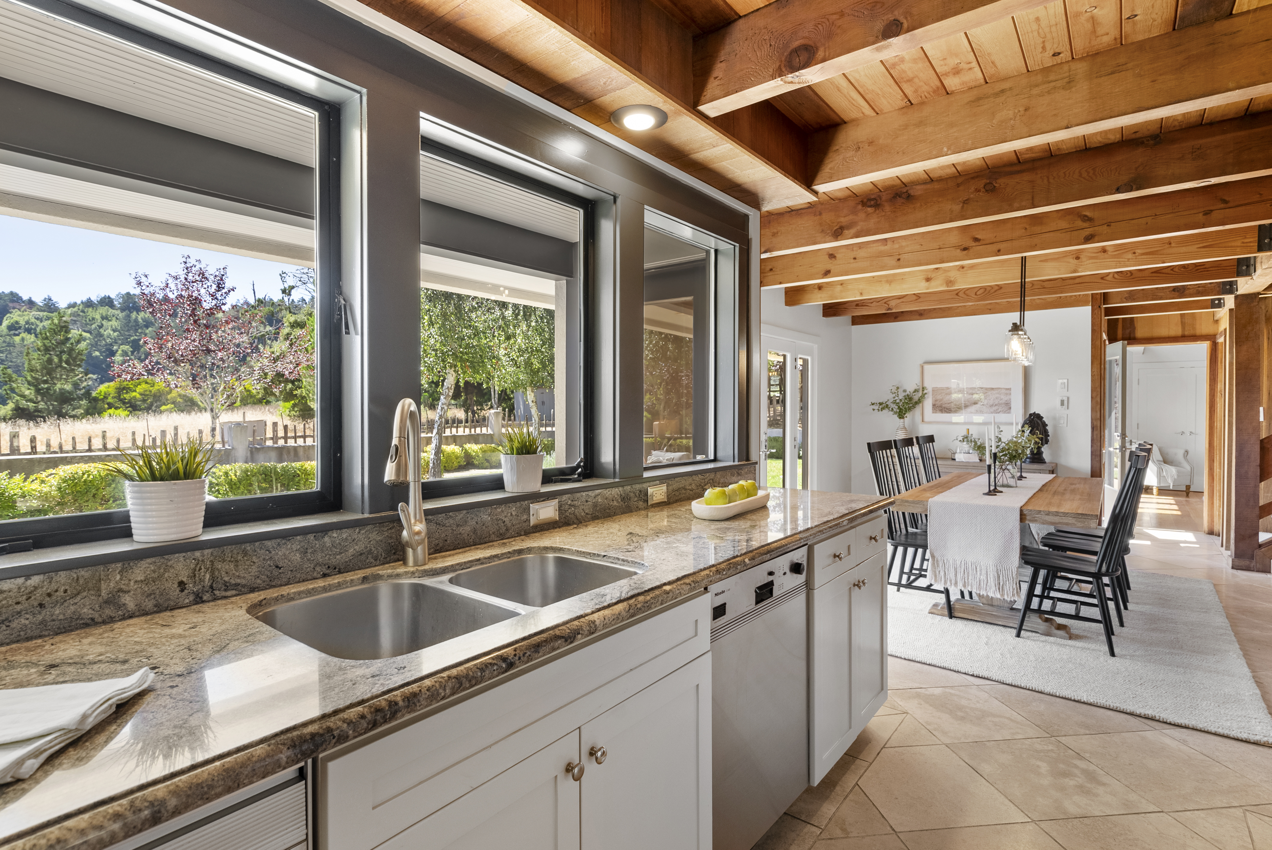 This bright, open kitchen features granite countertops, a double stainless steel sink, and white cabinetry, seamlessly connecting to an adjacent dining area. The space is defined by its exposed wood beam ceiling, creating a warm and inviting atmosphere. Large windows throughout provide ample natural light and scenic views of the lush outdoors.