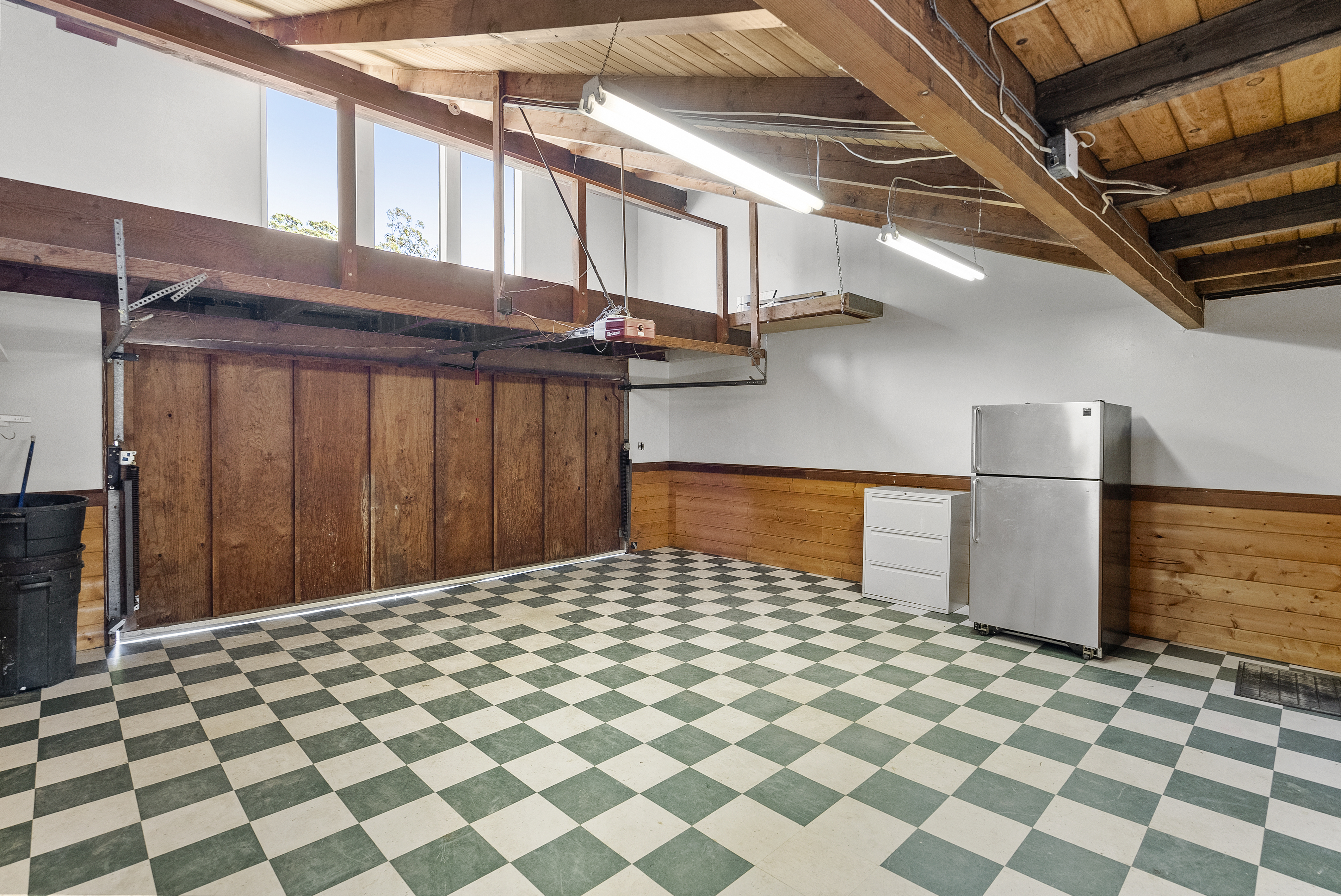 This interior garage space features a classic, rustic design with vaulted, exposed beam ceilings and checkerboard-patterned tile flooring. It includes a large wooden garage door, a prominent stainless steel refrigerator, and a small white filing cabinet, providing ample space for storage or workspace use. The presence of wood-paneled accents on the walls adds a warm, finished feel to the functional area.