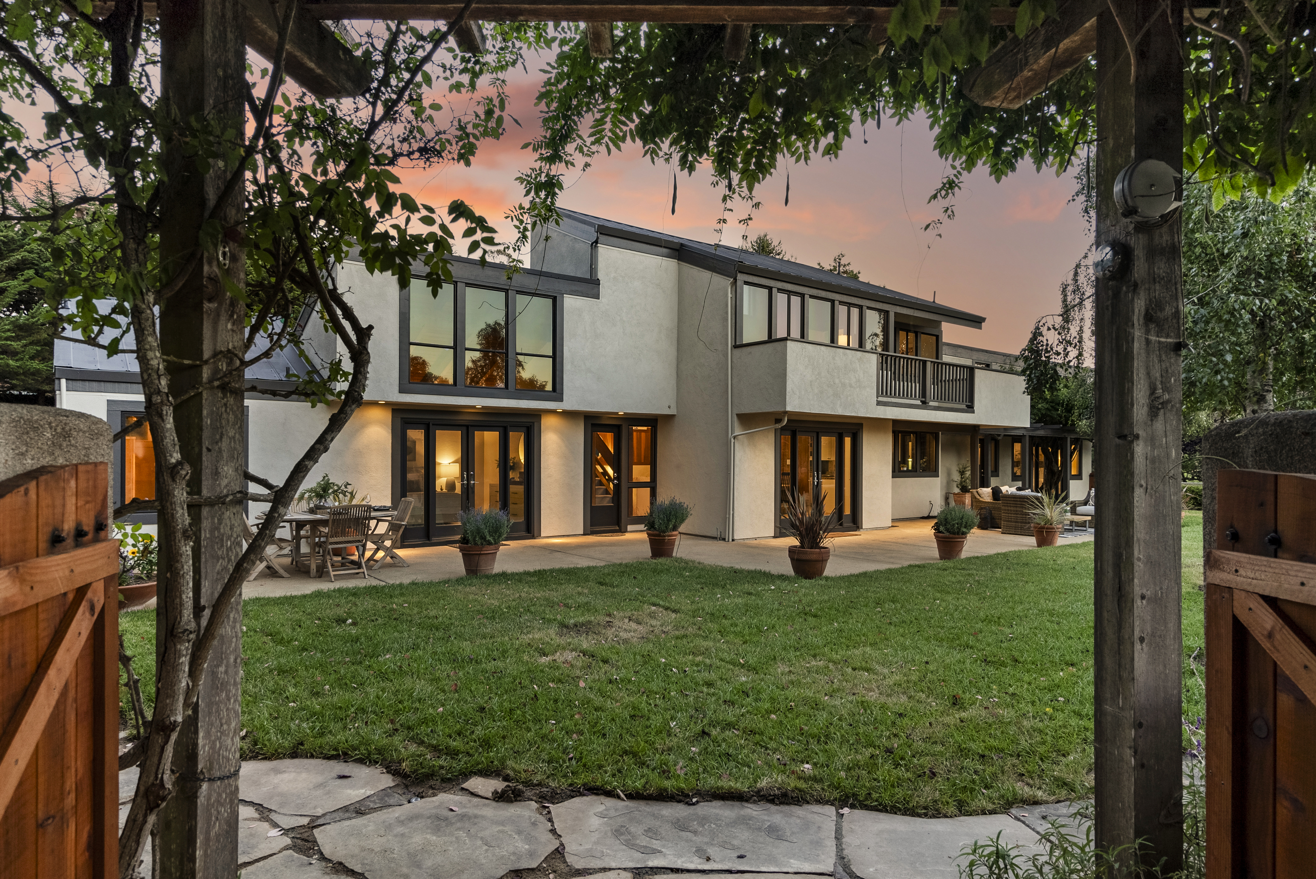This stunning contemporary two-story home is captured at dusk, showcasing an expansive concrete patio and a lush green lawn framed by romantic wooden pergola structures. The exterior features neutral stucco siding with updated dark-framed windows and doors, creating a sophisticated transition between indoor and outdoor living spaces. The property exudes a warm, inviting atmosphere, perfect for evening entertaining, with thoughtfully placed exterior lighting. 