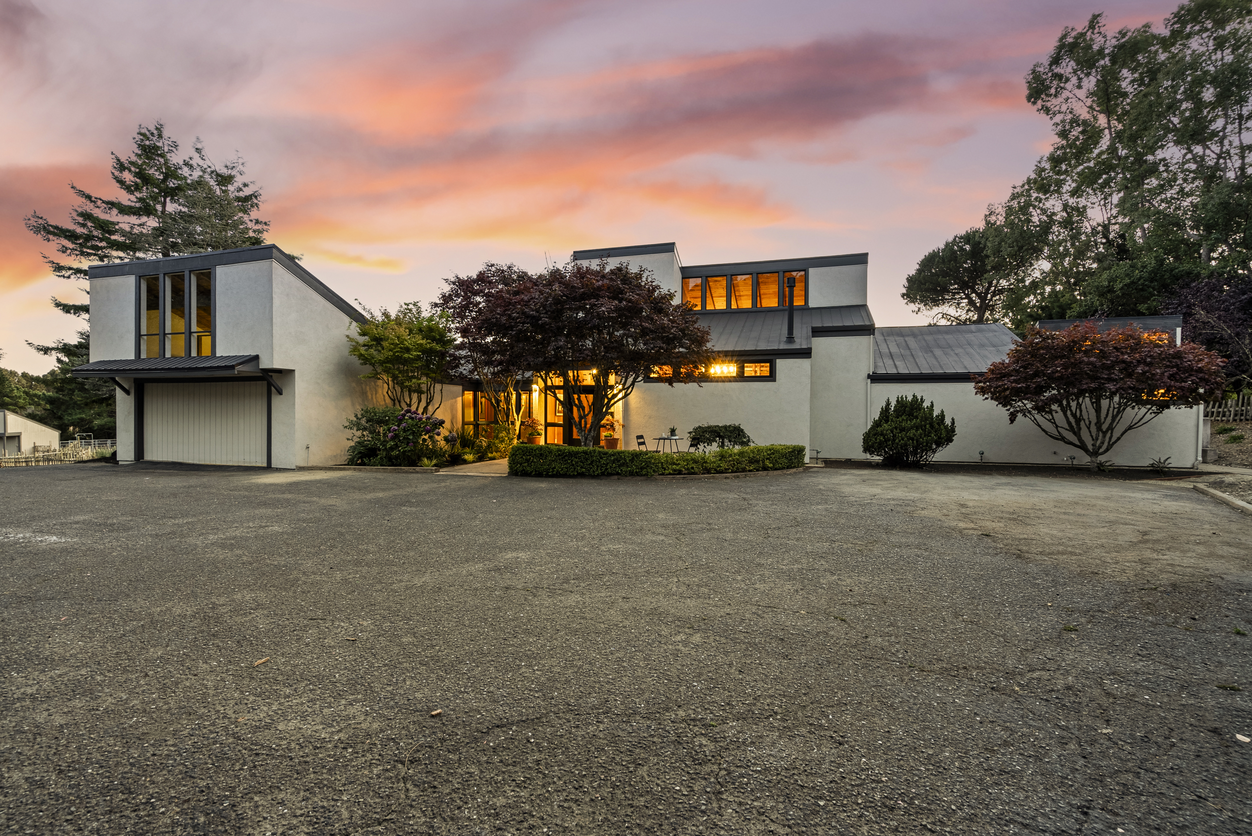 This stunning contemporary residence showcases a unique architectural design with dramatic angular rooflines and a crisp, light-colored stucco facade. Set against a breathtaking sunset sky, the property features beautiful mature landscaping, a prominent driveway, and warm interior lighting that invites a sense of modern elegance. The combination of geometric forms and natural scenery highlights the home's distinctive curb appeal.