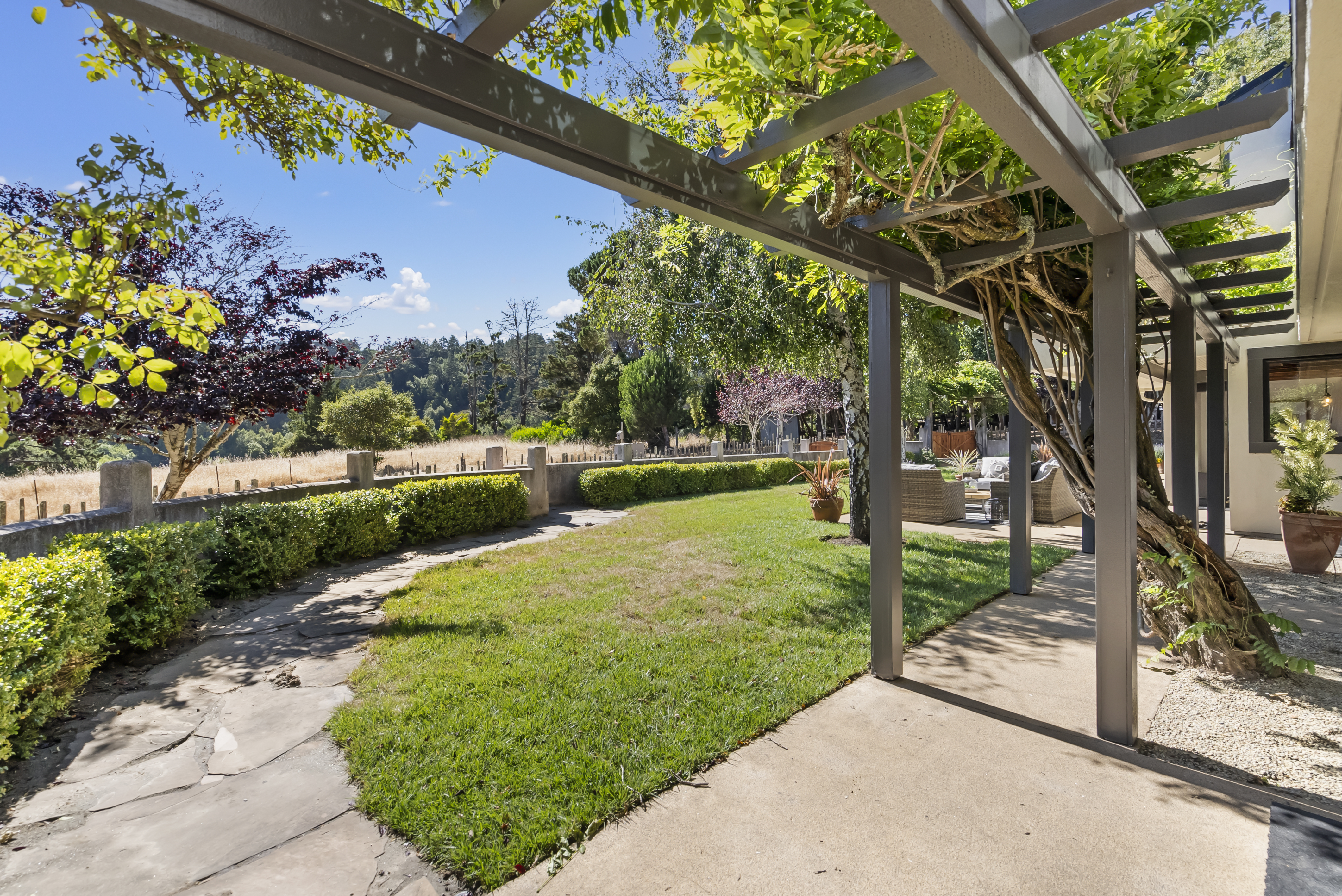 This outdoor living space features a large, covered concrete patio area shaded by an elegant wood pergola entwined with mature vines. The patio transitions seamlessly into a well-manicured lawn and a stone walking path, offering expansive views of a peaceful, rustic landscape. With comfortable seating arranged in the background, this area serves as an idyllic, private oasis perfect for relaxation or entertaining.