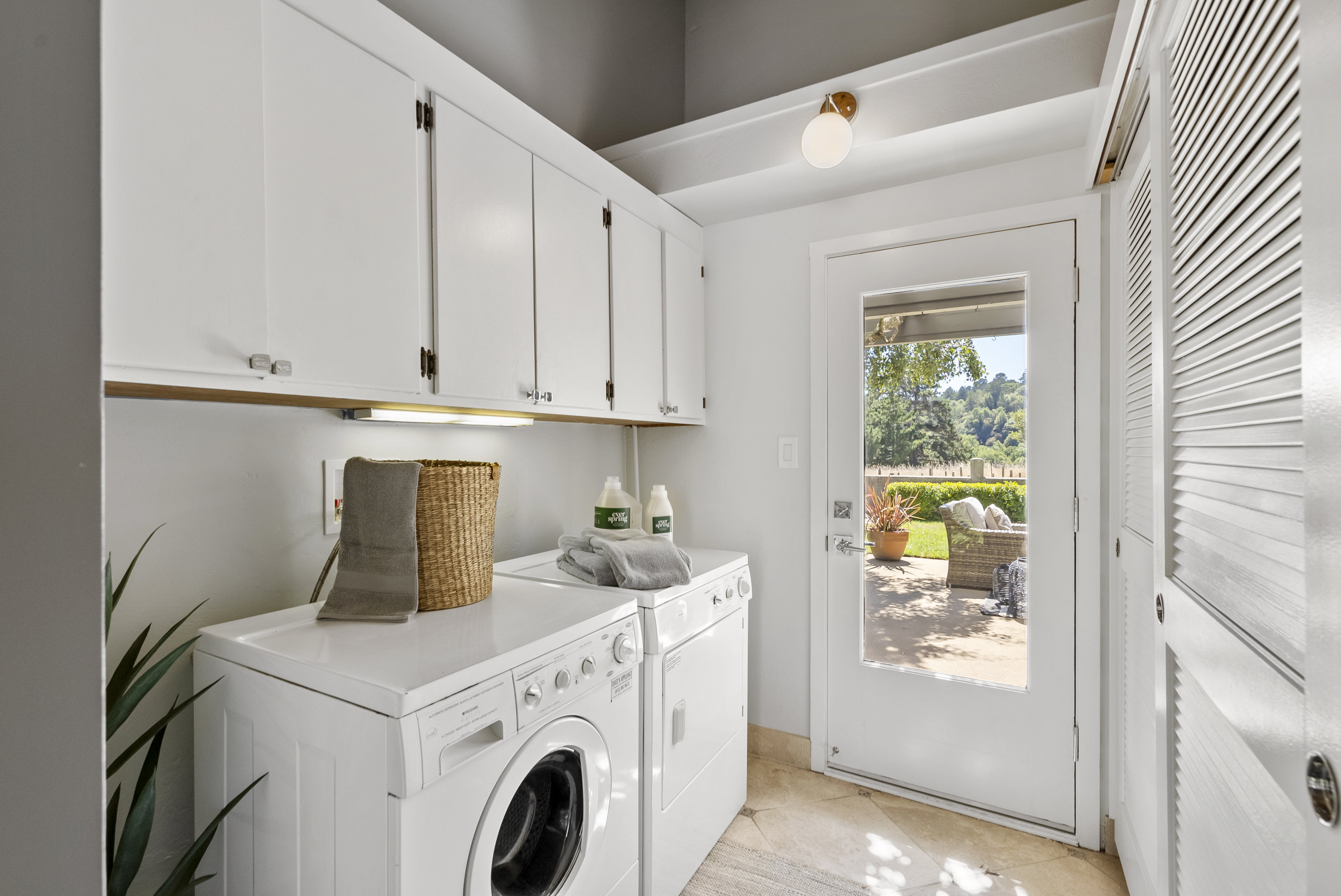 This bright laundry room features white cabinetry, high-efficiency appliances, and a convenient exterior door that opens to a patio. The space is efficiently designed for functionality, with a neutral color palette and clean lines that make it feel modern and organized. A large window on the door provides natural light and a pleasant view of the outdoor garden area.