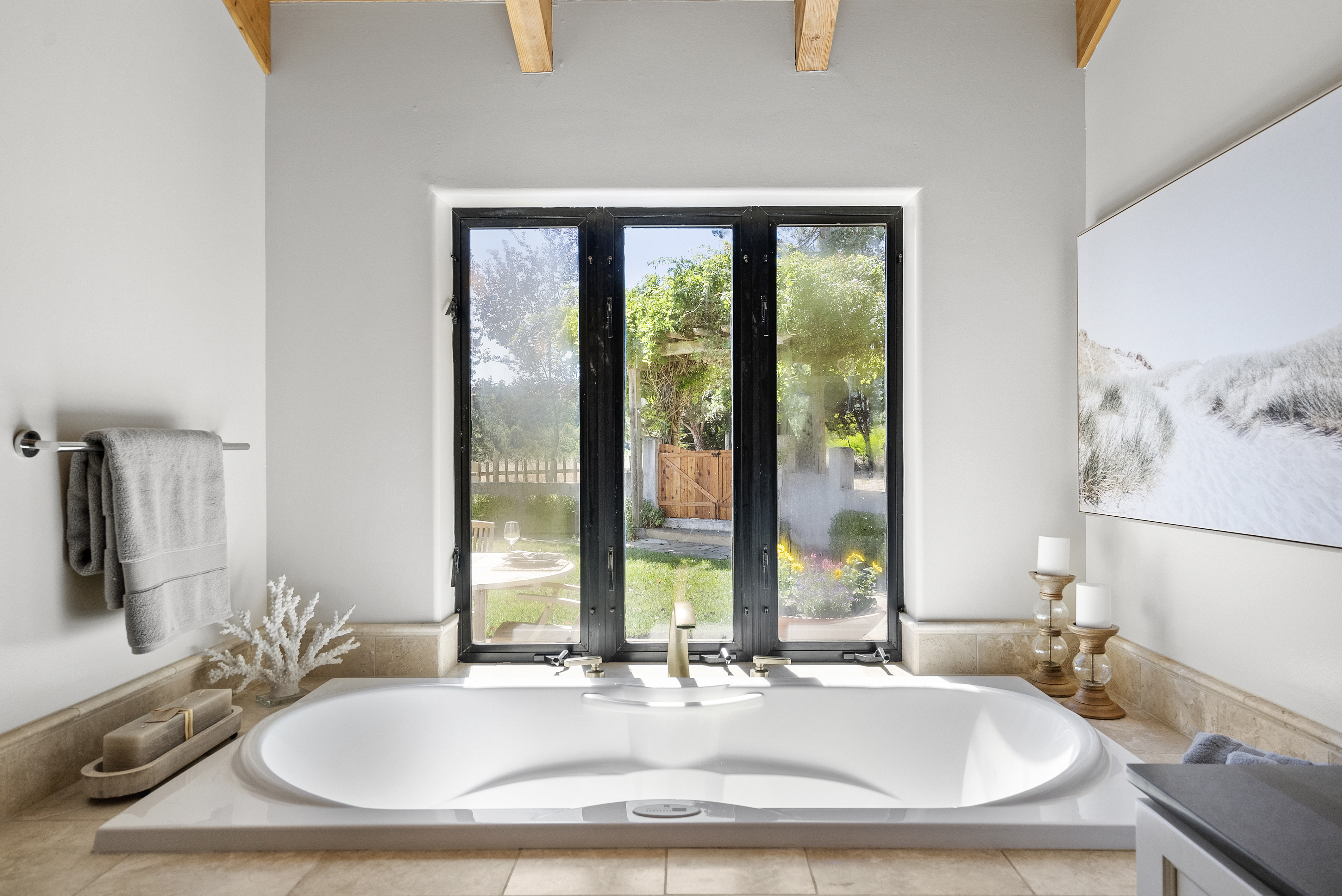 This serene primary bathroom features a deep soaking tub set against a large, black-framed picture window that offers a picturesque view of a lush garden. The space displays a neutral, elegant aesthetic with textured beige tiling, soft lighting, and tasteful decor like a white coral sculpture and candles. It provides a luxurious and tranquil atmosphere, perfect for a spa-like retreat within the home.