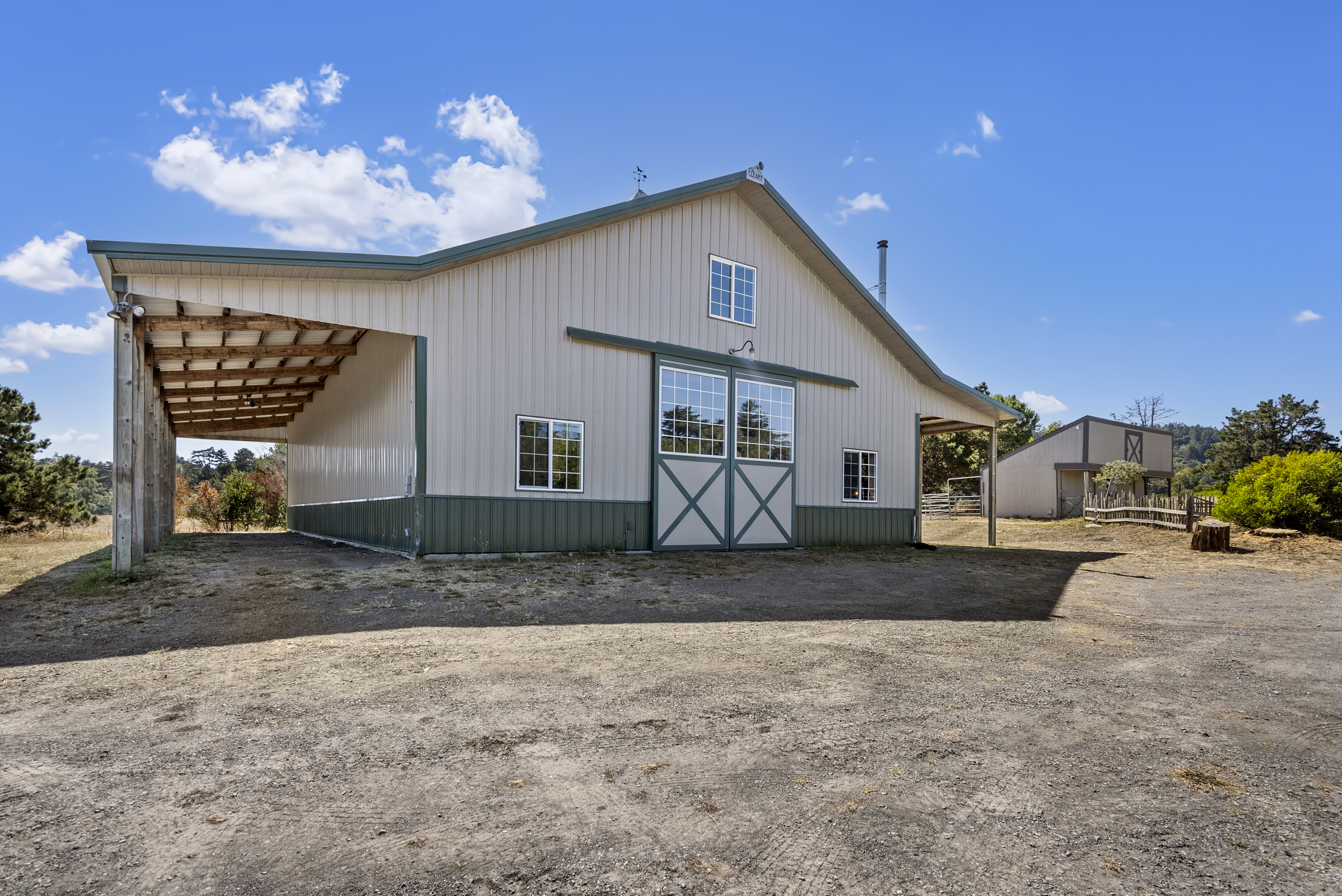 This image showcases a well-maintained, detached pole barn or workshop with a metal exterior, split between a beige upper section and a green lower wainscot. A large sliding barn door serves as the primary entrance, flanked by windows and an attached open-sided lean-to structure, creating a functional rural barn aesthetic. The property is set on a clear, gravel-surfaced lot under a vibrant blue sky, suggesting a spacious outdoor environment.