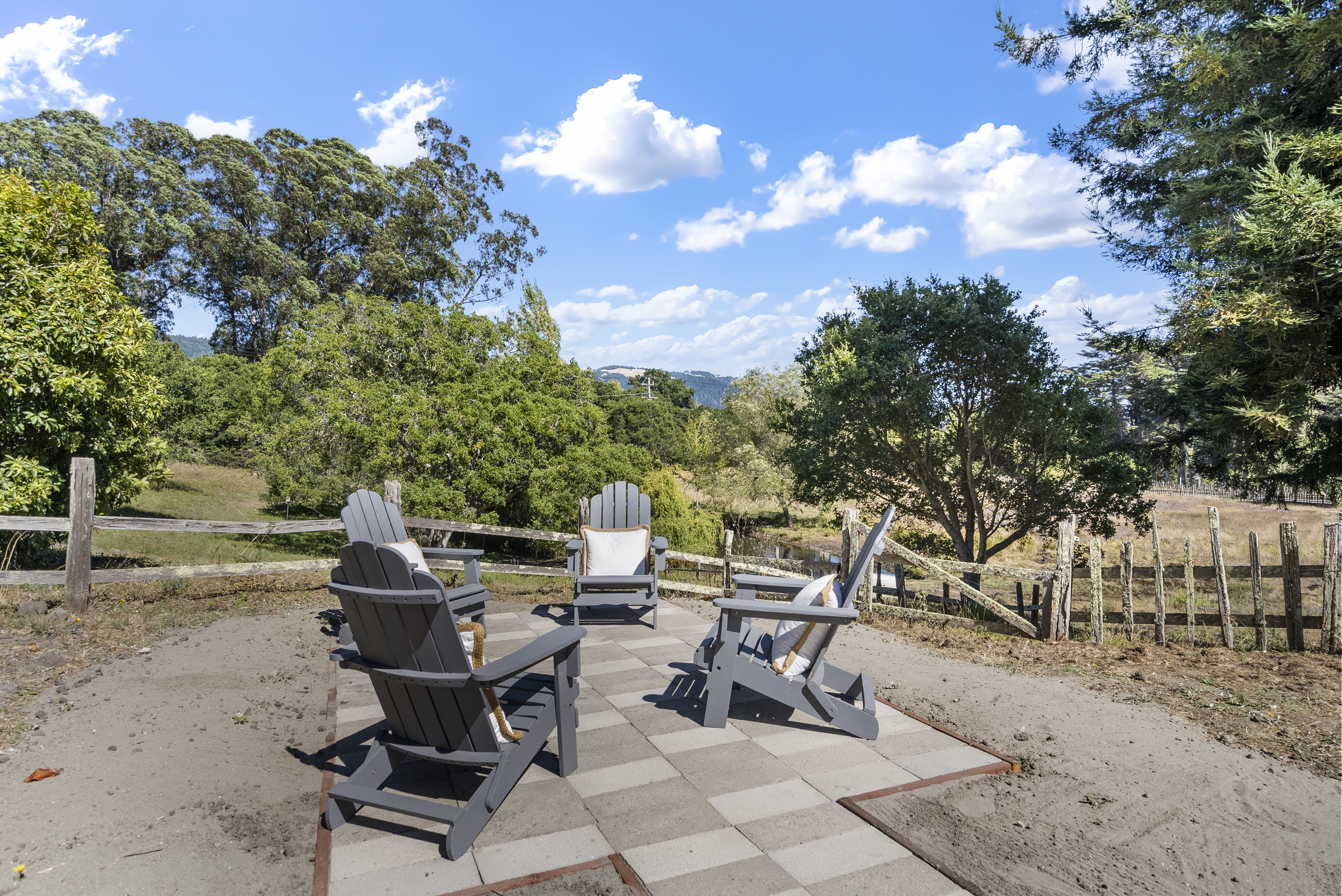 This outdoor patio space features a paved seating area with three gray Adirondack chairs, each accented with decorative pillows. It is positioned behind a rustic wooden fence in a expansive, park-like yard with mature trees and a scenic view of distant hills. The setting offers a peaceful and inviting space for relaxation in a natural, tranquil environment.