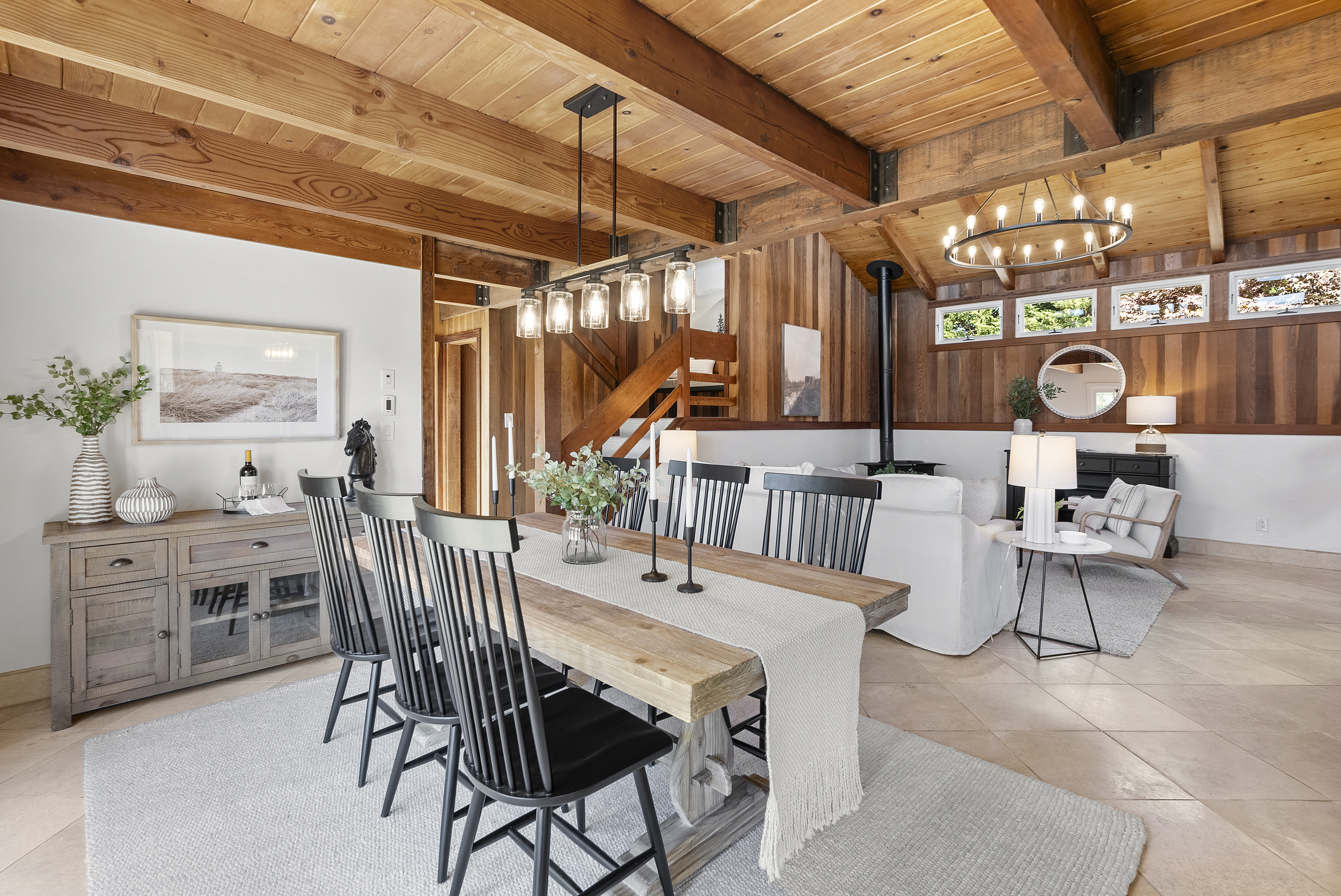 This spacious and inviting open-plan dining and living area features warm wood-beamed ceilings and a rustic-modern aesthetic. The dining space is anchored by a sturdy wooden table with black spindle-back chairs, while the connected living area highlights a cozy fireplace and large windows that bring in natural light. The combination of wood paneling and neutral tones creates a cohesive, high-end mountain-lodge feel throughout the home.