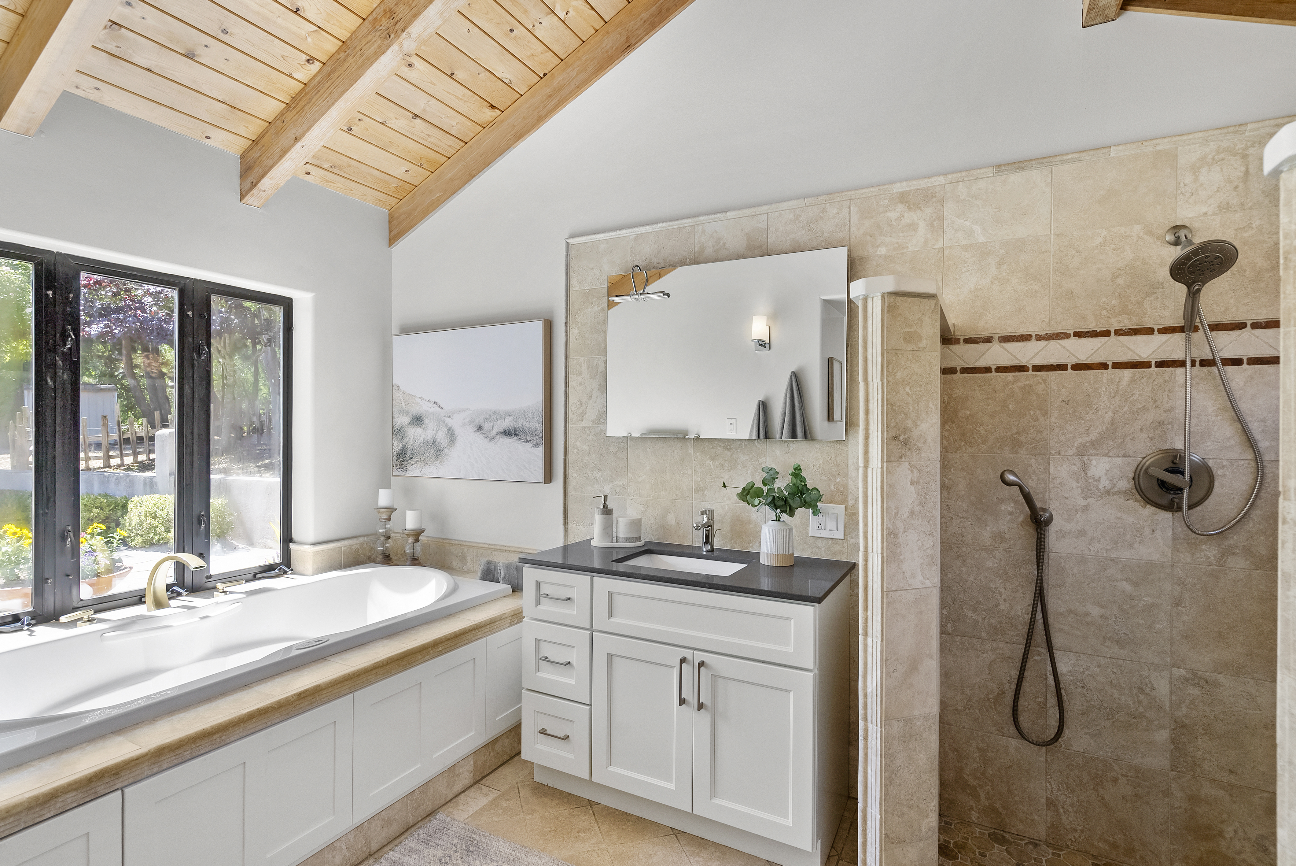 This elegant primary bathroom features a vaulted ceiling with exposed wooden beams and a large soaking tub positioned beneath a window. The space is equipped with a modern white vanity, a dark countertop, and an adjacent walk-in shower finished with neutral-toned stone tiling. The combination of warm wood tones and natural stone creates a bright, inviting spa-like atmosphere.