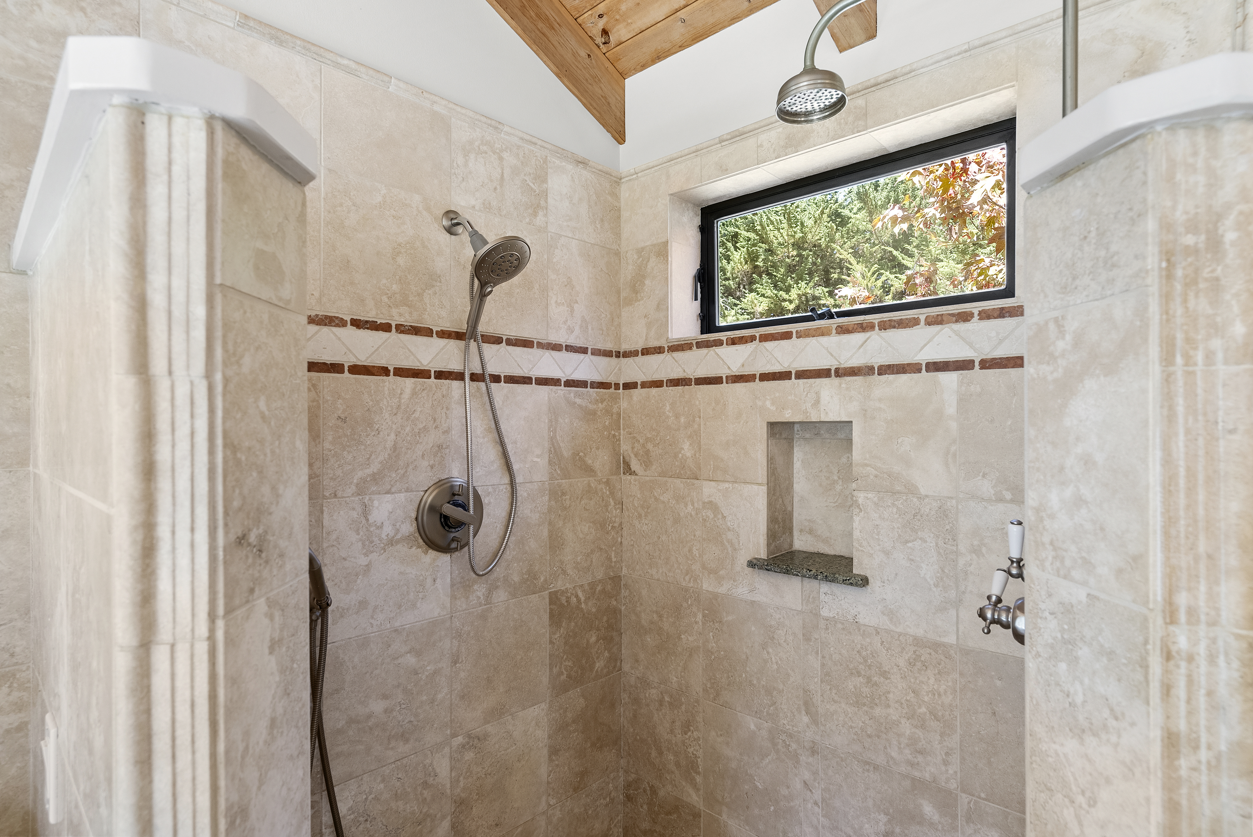 This primary bathroom shower features neutral stone tiling with an elegant decorative accent border. It includes a walk-in design with a rainfall showerhead, a separate handheld shower nozzle, built-in shelving, and a window allowing natural light into the space, all framed by rustic wood ceiling accents.