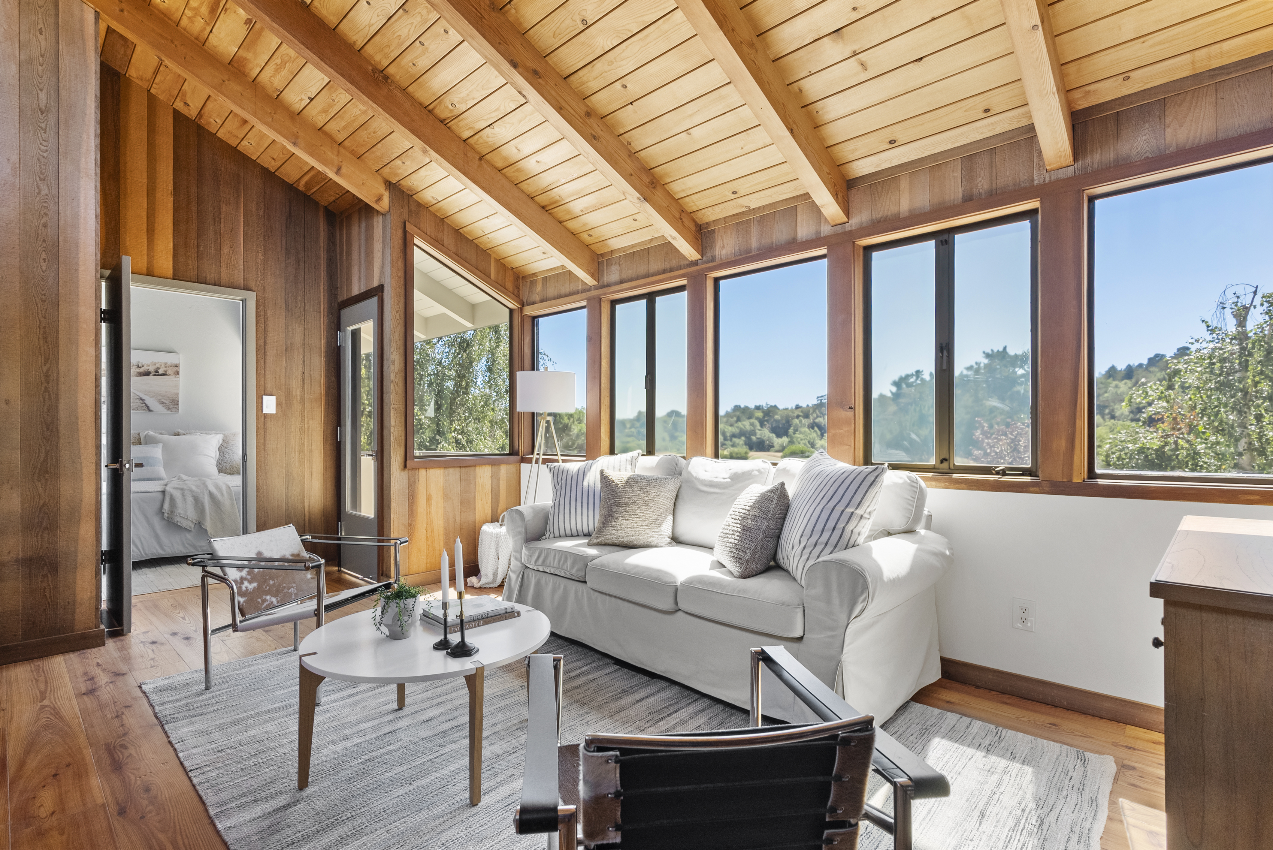 This cozy living room features warm wood-paneled walls and a matching vaulted ceiling with exposed beams. The space is furnished with a comfortable white sofa and contemporary accent chairs arranged around a circular coffee table, all set against a view of the outdoors through a series of large windows. The overall aesthetic is rustic yet modern, offering a bright and inviting atmosphere.