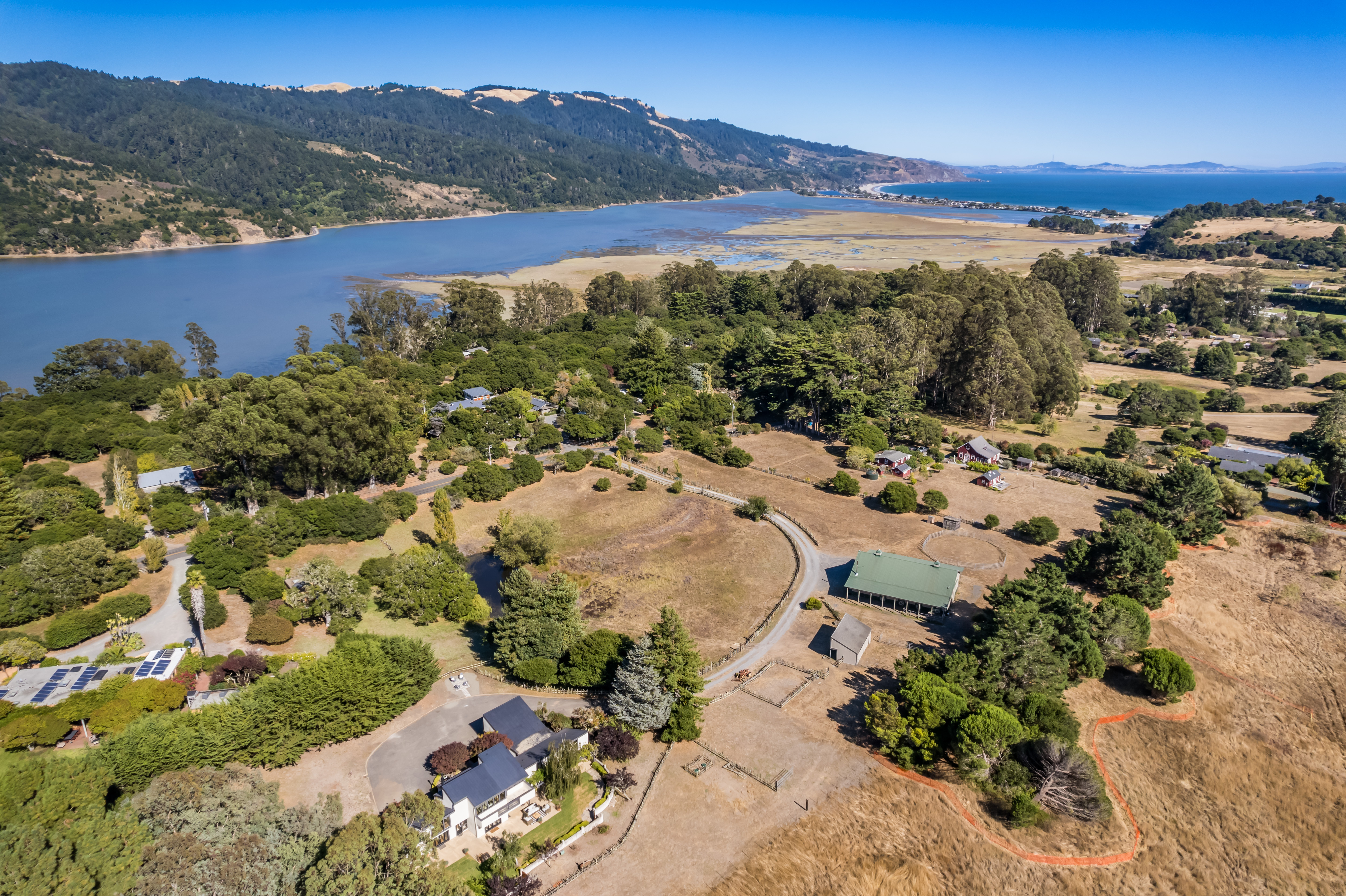 This panoramic aerial view showcases a sprawling rural property nestled between lush hills and a picturesque coastline. The estate features multiple structures, including a primary residence with solar panels, a large barn, and open fields, all surrounded by extensive greenery and natural landscapes. Its expansive layout and serene coastal proximity highlight the property's immense potential for privacy and scenic living.