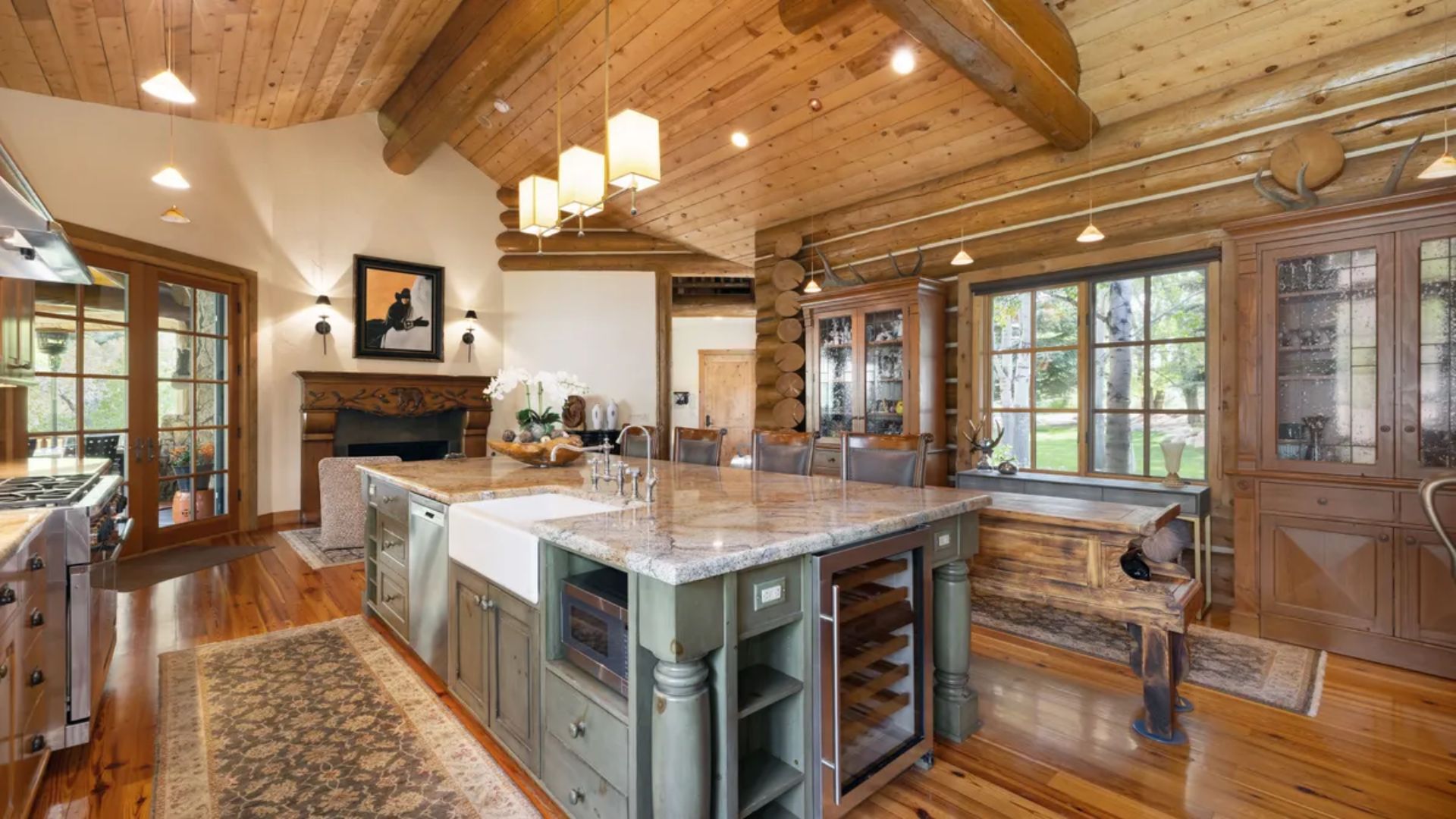 This rustic, lodge-style kitchen features a large central island with an apron-front sink, wine cooler, and built-in microwave. The space is highlighted by warm wood paneled ceilings, stone flooring, and a dining bench situated near a large window. A stone fireplace and a glass-front display cabinet add to the cozy yet luxurious atmosphere, making it a perfect space for both cooking and entertaining.