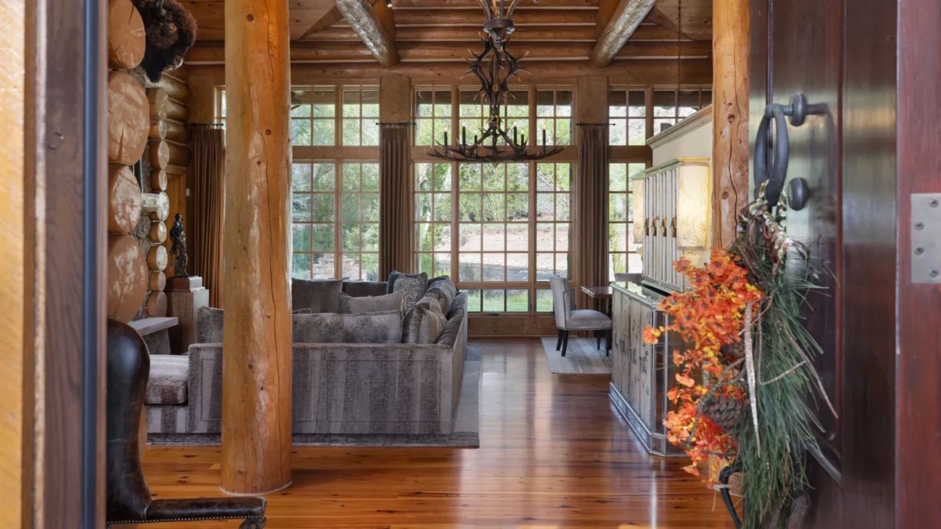 This rustic living room features massive log support columns, exposed timber ceiling beams, and polished wood flooring, creating a warm and authentic cabin atmosphere. A large, plush sofa serves as the center point, complemented by floor-to-ceiling windows that provide an expansive view of the outdoors. The space is anchored by a unique antler chandelier, enhancing the luxurious, mountain-lodge aesthetic throughout the room.