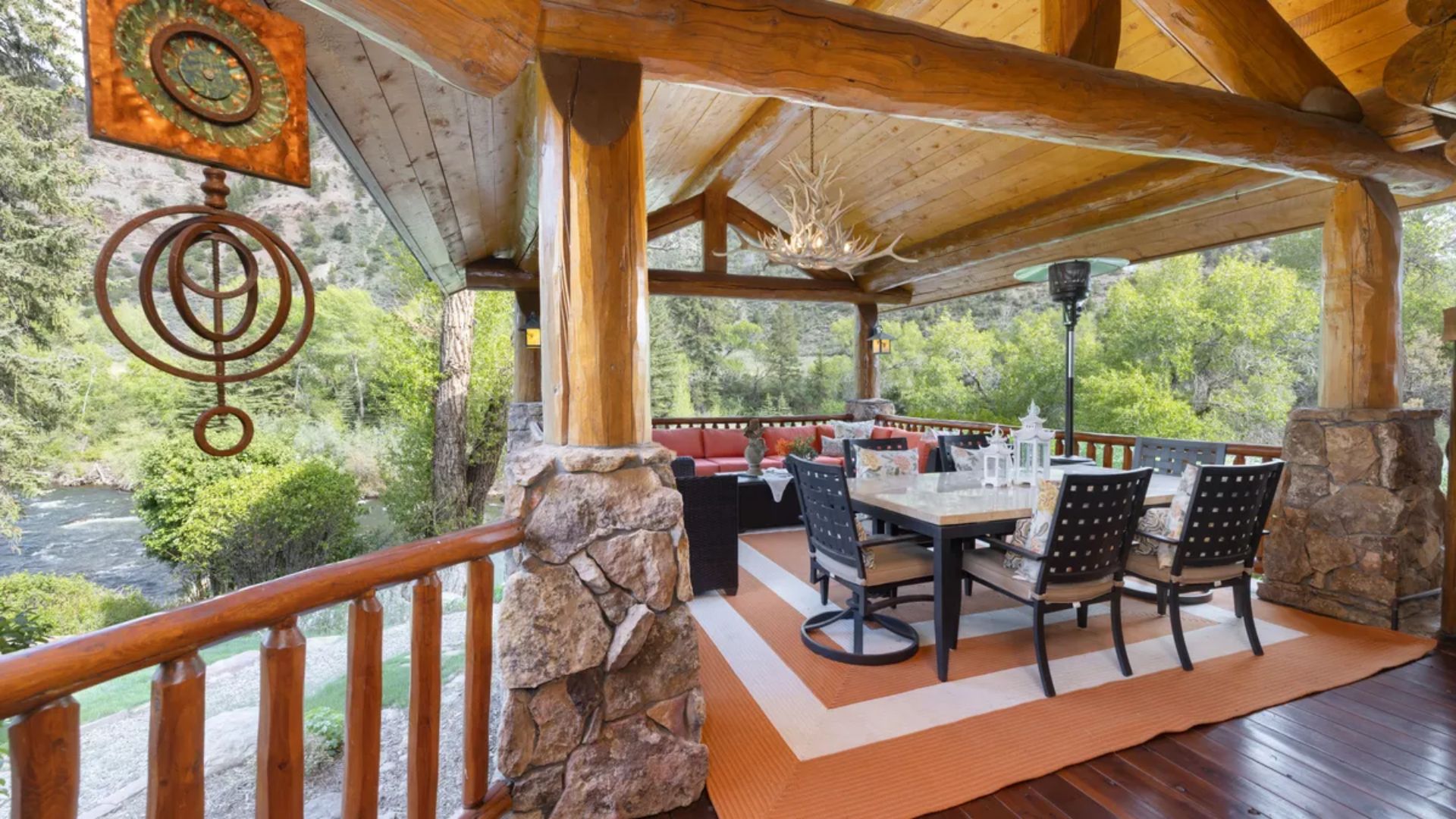 This spacious, covered outdoor patio features a rustic, log-cabin aesthetic with exposed wooden beams, stone support pillars, and a ceiling-mounted antler chandelier. The area is furnished with a dining table and comfortable lounge seating, making it an ideal space for entertaining while overlooking a scenic, tree-lined river view. The space combines durability with luxury, offering a tranquil retreat that seamlessly blends with the natural surroundings.