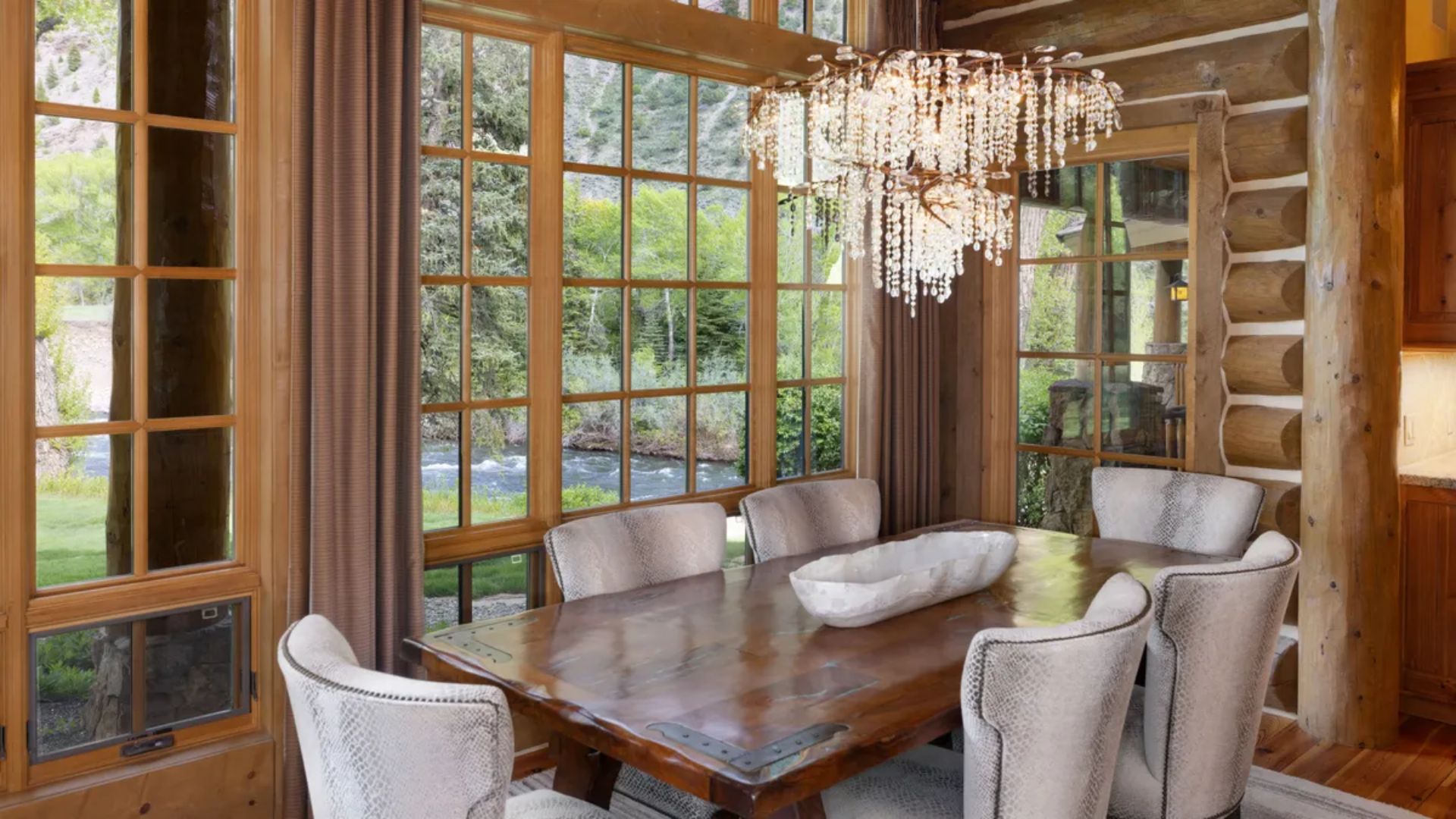 This rustic, lodge-style dining room features a solid wood dining table accented with metallic details, surrounded by textured, neutral-toned upholstered chairs. A large, ornate crystal chandelier hangs above the table, while expansive grid-pane windows provide natural light and picturesque views of the adjacent river and landscape. The space harmoniously blends luxury fixtures with the warm, rugged aesthetic of wood-logged walls.
