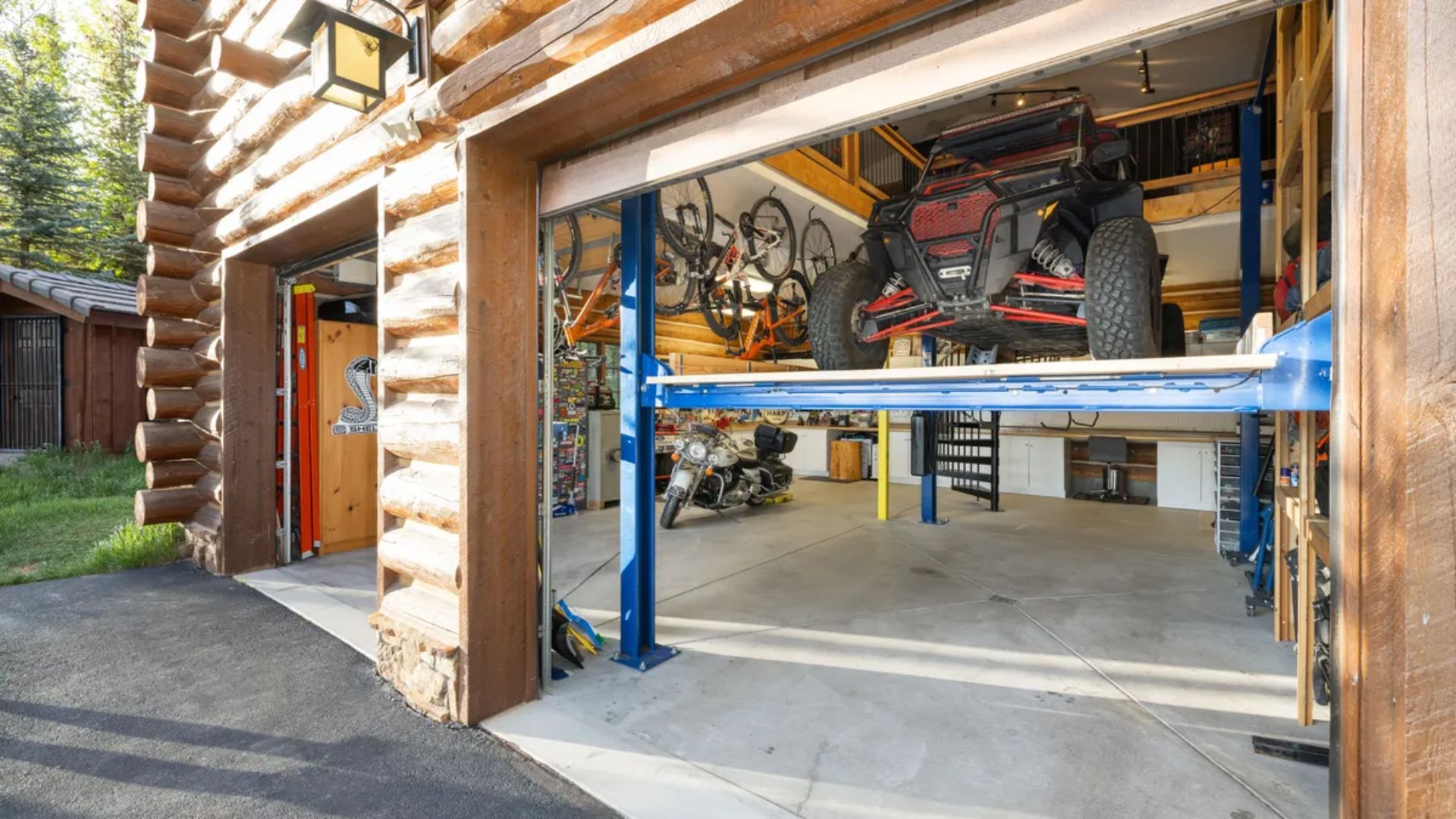 This spacious and rustic-style garage features distinctive log exterior siding and an open bay showcasing an elevated storage system with a UTV. The interior offers a practical workspace with concrete flooring, a motorcycle, and various wall-mounted items, emphasizing versatile storage solutions. It provides an impressive blend of rugged exterior aesthetic and functional utility space suitable for car enthusiasts or hobbyists.