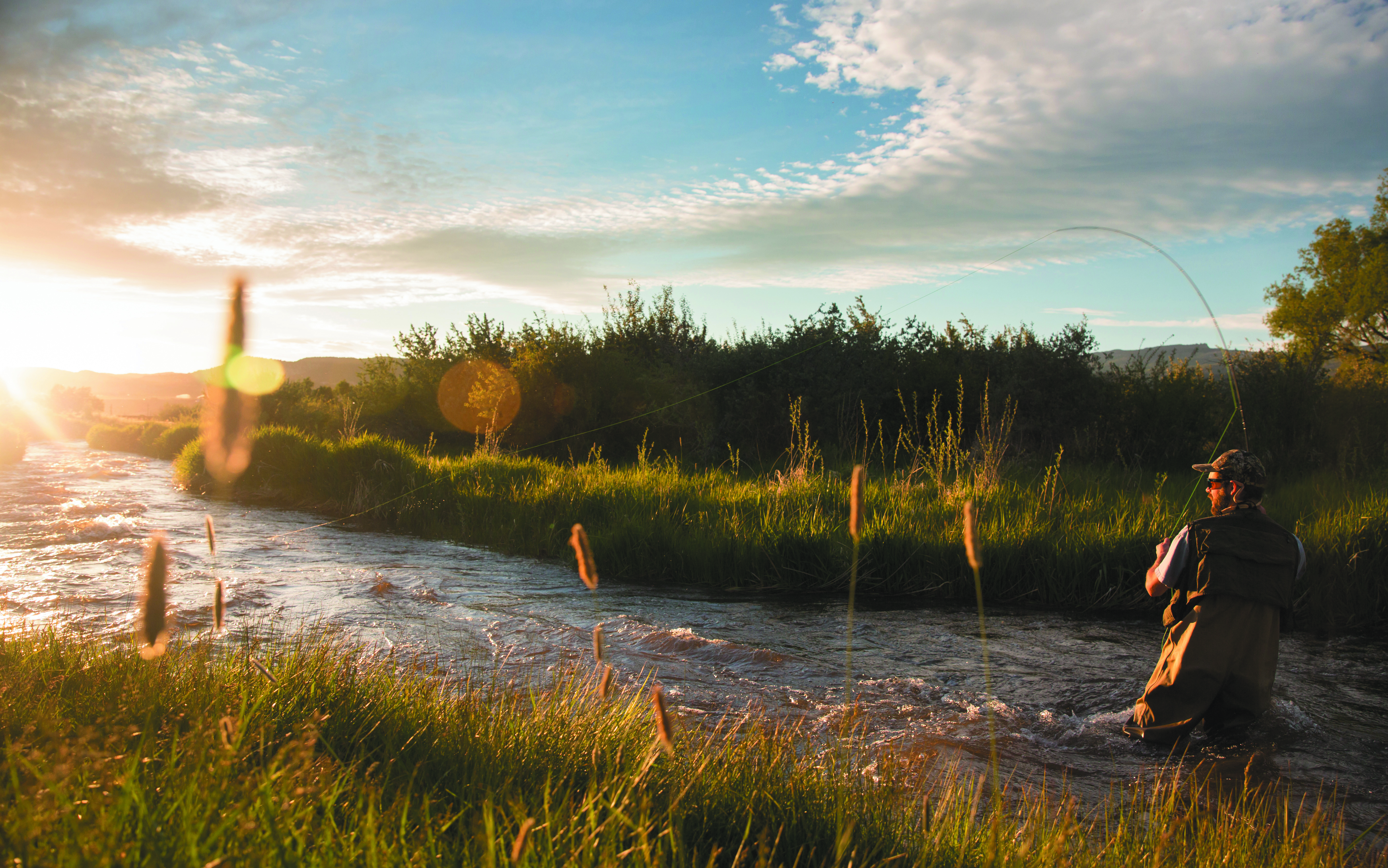 This scenic image features a fly fisherman casting his line in a peaceful river situated within a rural, park-like landscape. The golden hour lighting highlights the lush green grasses, tranquil water, and open natural setting, suggesting excellent opportunities for outdoor recreation and waterfront living. This imagery would be ideal for highlighting high-quality community amenities in a luxury development or outdoor-focused residential community.