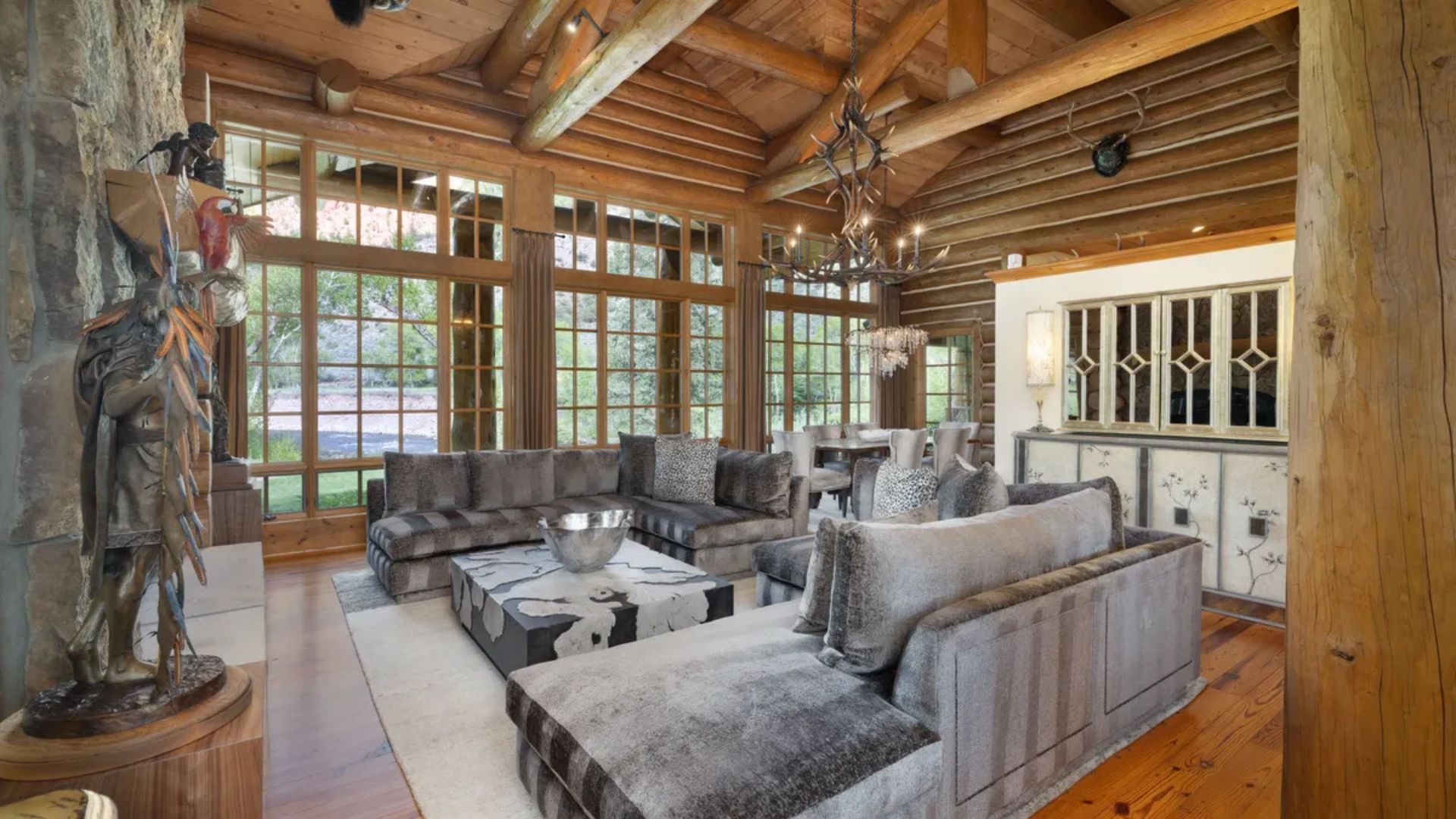 This rustic living room features an impressive vaulted ceiling with exposed log beams and a grand antler chandelier, emphasizing a cozy cabin aesthetic. The space is anchored by a large, contemporary stone fireplace and expansive floor-to-ceiling windows that provide abundant natural light and scenic views. Plush, oversized sectional seating surrounds a modern coffee table, creating a luxurious yet comfortable atmosphere perfect for relaxing.