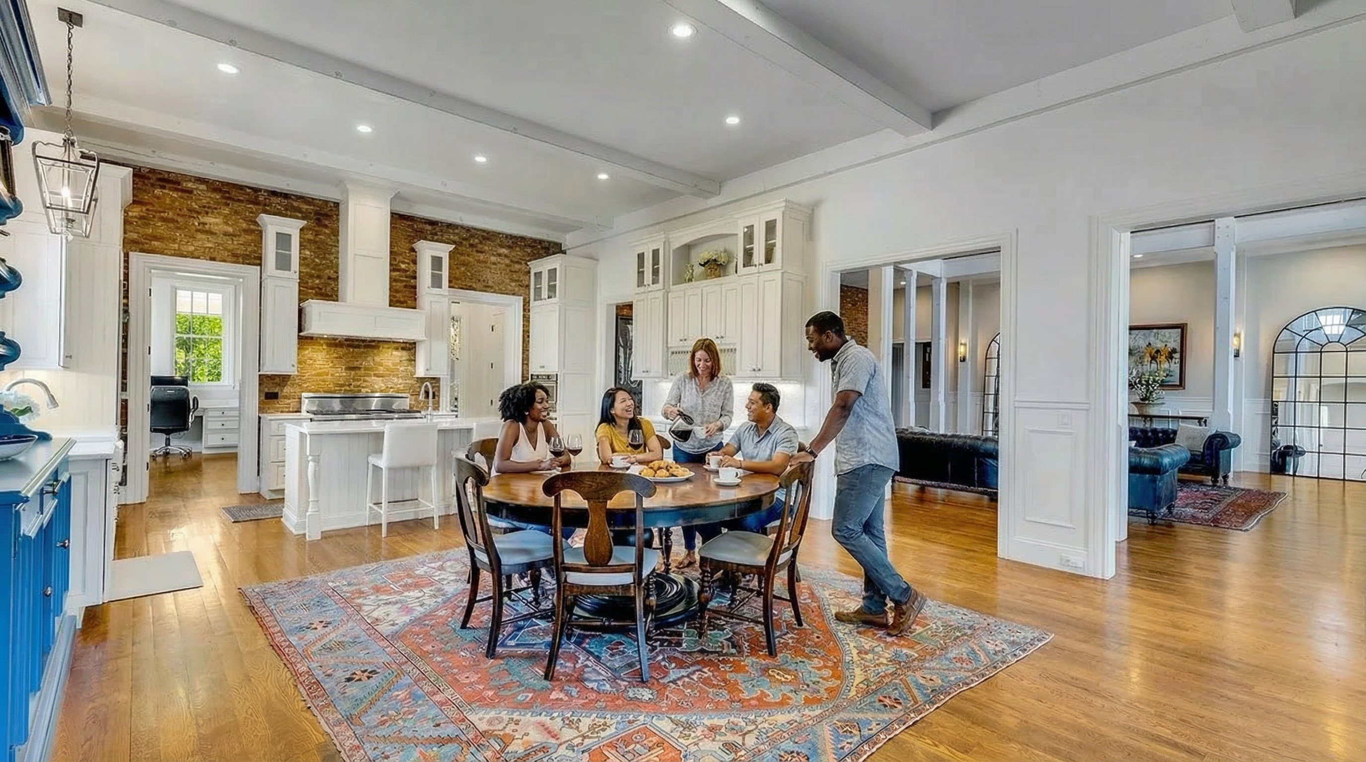 This bright, open-plan kitchen and dining area features elegant white cabinetry, a striking exposed brick backsplash, and warm hardwood flooring. The space is anchored by a large circular dining table, creating an inviting atmosphere for social interaction, and connects seamlessly to other rooms through open doorways. The high-end finishes and architectural charm contribute to a sophisticated, modern-traditional living environment.