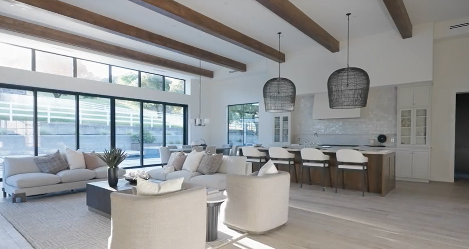 This spacious, open-concept living area seamlessly connects a comfortable lounge space with a modern, high-end kitchen. Key features include vaulted ceilings with exposed wood beams, expansive floor-to-ceiling windows that provide natural light, and a stylish neutral color palette. The room radiates a sophisticated, coastal modern aesthetic, ideal for both relaxed family living and elegant entertaining.