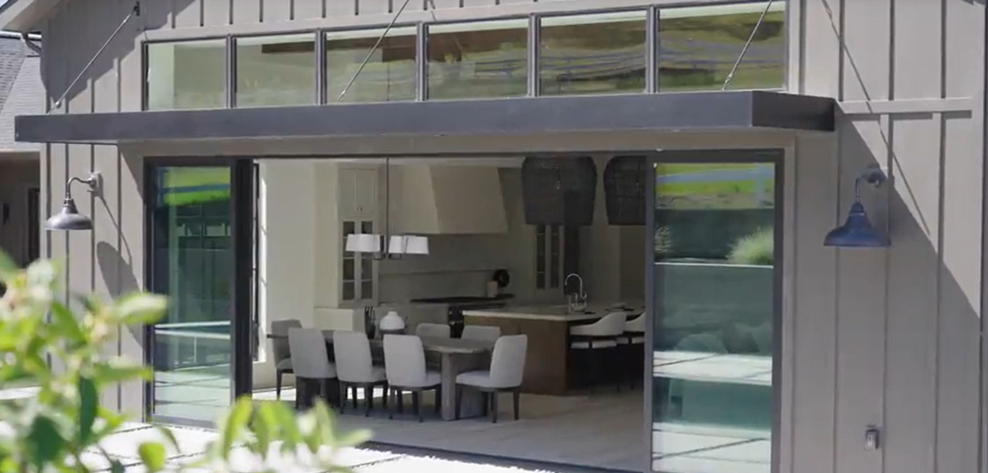 This rear view showcasing an indoor-outdoor living concept features a seamless transition between a modern kitchen and dining space and the outdoors. Large, dark-framed sliding glass doors open wide, framed by a striking black awning and industrial-style gooseneck wall lights, reflecting a contemporary farmhouse aesthetic.