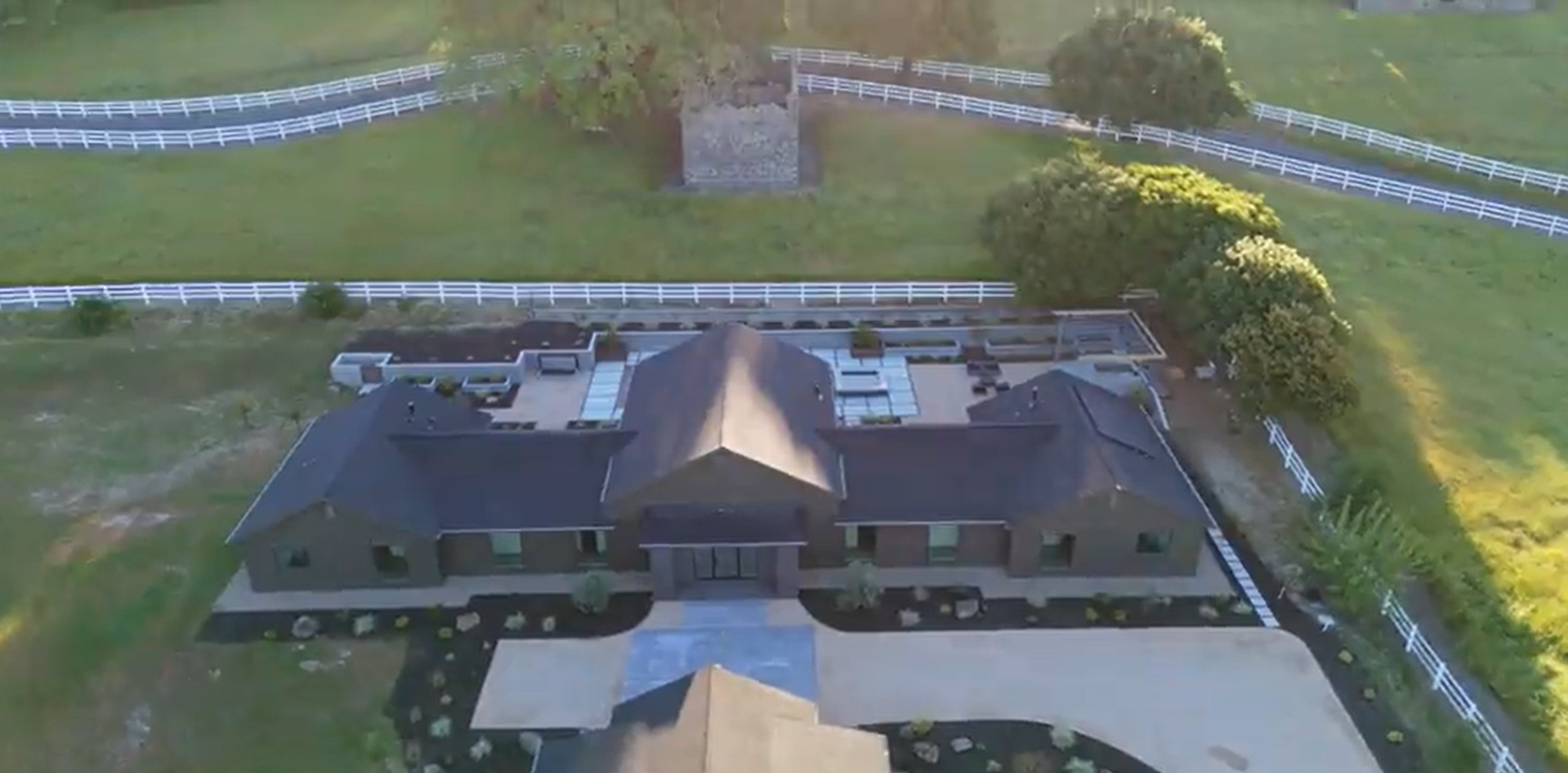 This stunning aerial view captures a contemporary, dark-toned sprawling residence nestled within a vast, rural estate. The property features extensive white fencing defining the rolling green pastures, a stone structure ruin in the background, and a well-manicured patio area. The home's symmetrical design and sophisticated landscaping contribute to a serene and expansive country living atmosphere.