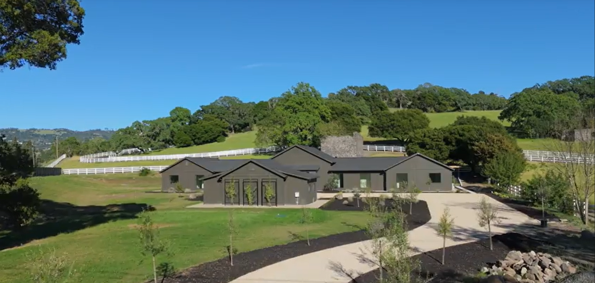 This image showcases a modern, monochromatic dark gray farmhouse set against a picturesque rolling landscape. The property features a wide, paved driveway leading to the home, surrounded by neatly manicured landscaping, green grass, and white ranch-style fencing. The architectural design is clean and minimalist, offering a striking contrast against the bright blue sky and natural surroundings.