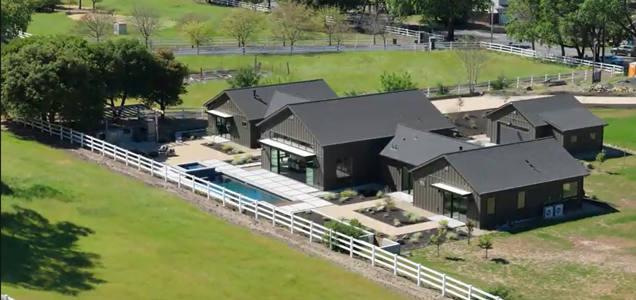 This stunning contemporary residence features dark siding and a modern aesthetic, set within a spacious, pastoral landscape. The property includes a sleek rectangular swimming pool, expansive paved patios, and multiple connected structures, all bordered by a classic white ranch-style fence. The elevated perspective highlights the seamless integration between the modern home design and its tranquil, rural surroundings.