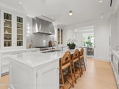This is a bright and spacious kitchen featuring white cabinetry, marble countertops and backsplash, and stainless steel appliances, including a large range hood. A center island provides ample workspace and includes bar seating. The kitchen connects to a dining area or breakfast nook, creating an open and inviting space for cooking and entertaining.