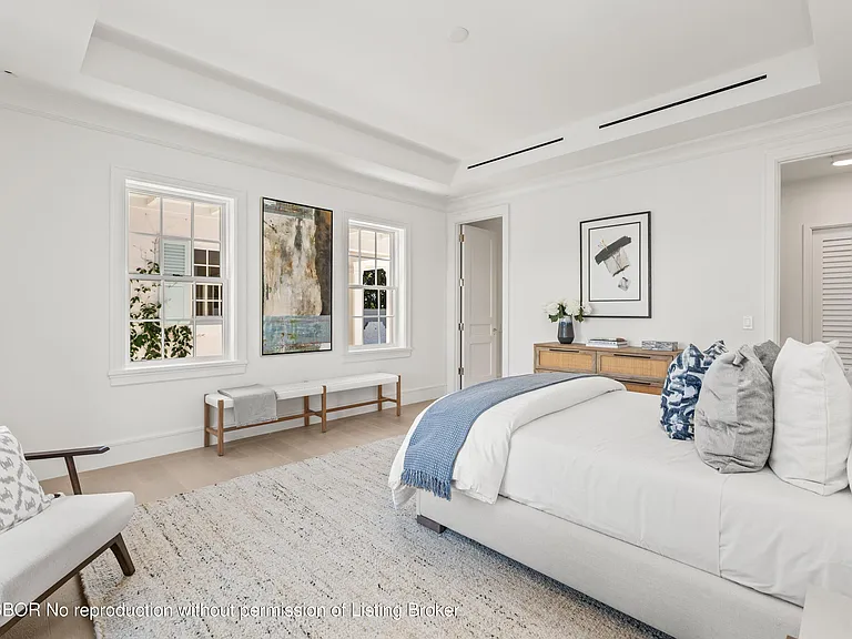 This is a primary bedroom featuring a large bed with white bedding and a blue throw. The room has ample natural light from the windows and artwork hanging on the wall. The overall aesthetic is clean, modern, and inviting, contributing to a serene and relaxing atmosphere.