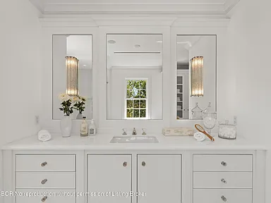 This is a well-lit primary bathroom featuring a white vanity with ample storage space. The vanity is topped with a bright white countertop and stylish fixtures. Three mirrors above the counter add to the room's brightness and create a sense of spaciousness.