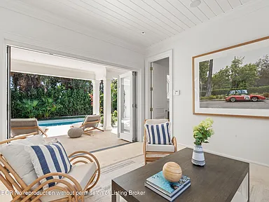 This image showcases a bright and stylish office/study area with direct access to the outdoor pool area. The room features neutral tones, comfortable seating, and a large photograph on the wall that adds character. The folding doors leading to the pool area create an indoor-outdoor living experience.