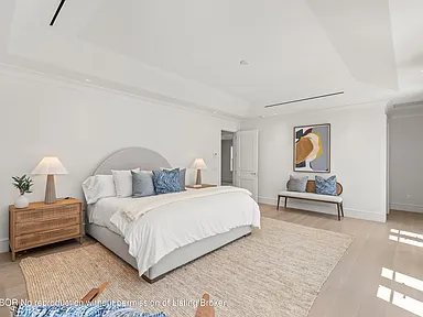 This is a bright and airy primary bedroom featuring a neutral color palette. The room showcases a king-size bed with light-colored linens and decorative pillows, complemented by matching bedside tables and lamps. Natural light floods the room, creating a serene and inviting atmosphere conducive to relaxation.