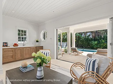 This interior shot captures a bright living room leading to an outdoor patio and pool area. The room features white vaulted ceilings, built-in cabinetry, and comfortable wicker seating adorned with striped cushions. Large glass doors open to the patio, seamlessly blending indoor and outdoor living spaces.
