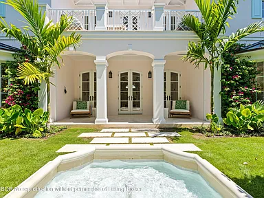 The image showcases a luxurious backyard featuring a spa or small pool in the foreground, surrounded by lush greenery and a well-manicured lawn. A covered patio area with seating is visible in the background, attached to a pristine white building with column supports. Tropical plants enhance the ambiance of this outdoor space, suggesting a premium property in a warm climate.
