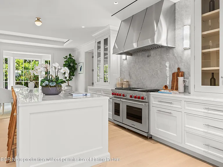 This is a bright and modern kitchen featuring white cabinetry and a large island with a marble countertop. The stainless steel range hood and professional-grade oven provide a sleek and functional focal point. The large windows and open layout create an inviting and spacious cooking environment.